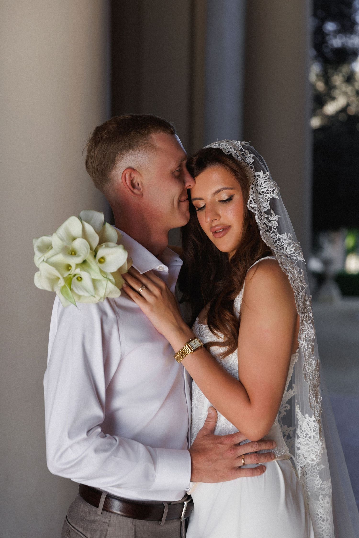 Bride and groom embrace tenderly, bride holding white calla lily bouquet, wearing lace veil and wedding dress.