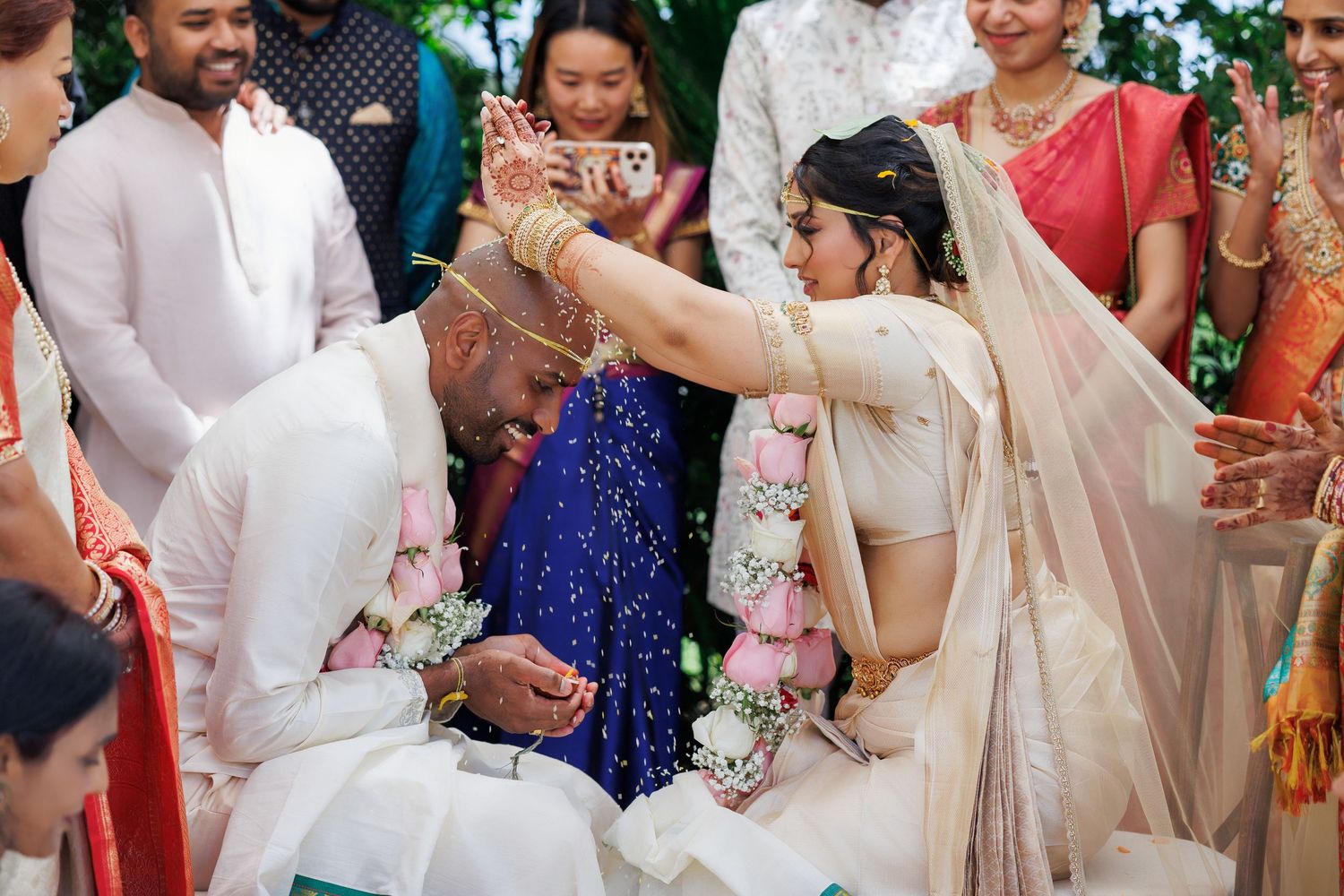Bride performs traditional Hindu wedding ritual pouring rice on groom during colorful outdoor ceremony with guests.