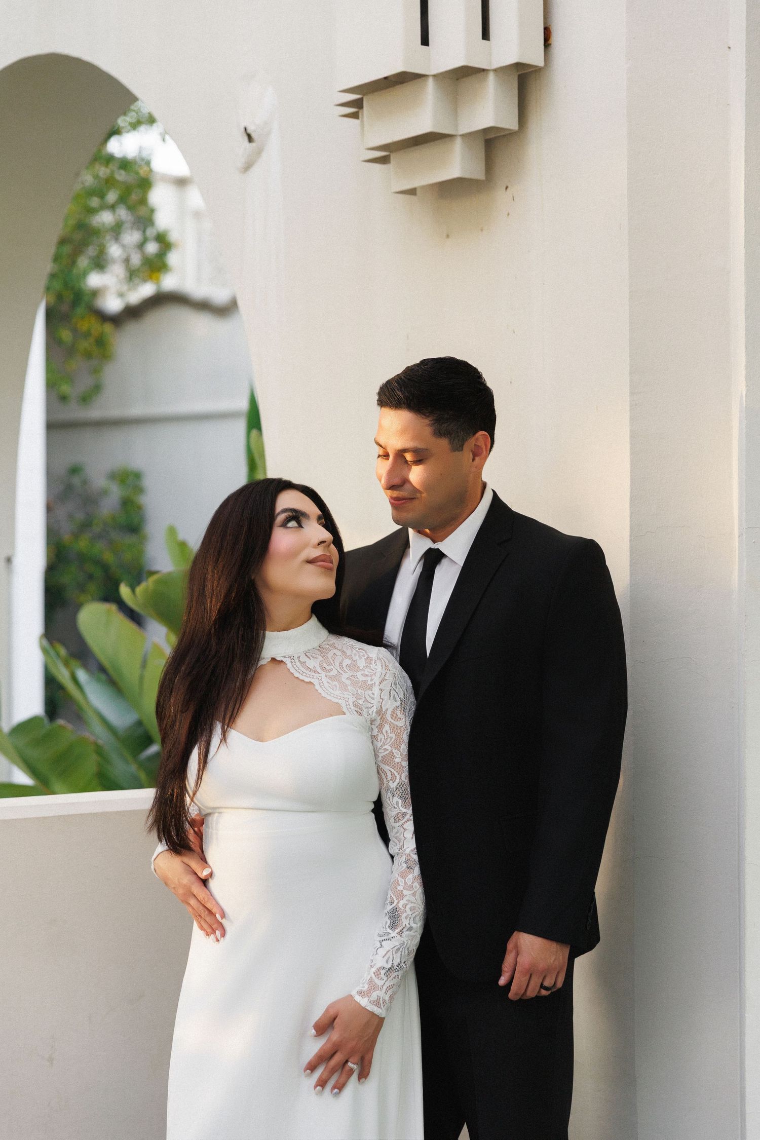 A couple in wedding attire poses by a white building, the bride in lace dress gazing up at the groom in a black suit.