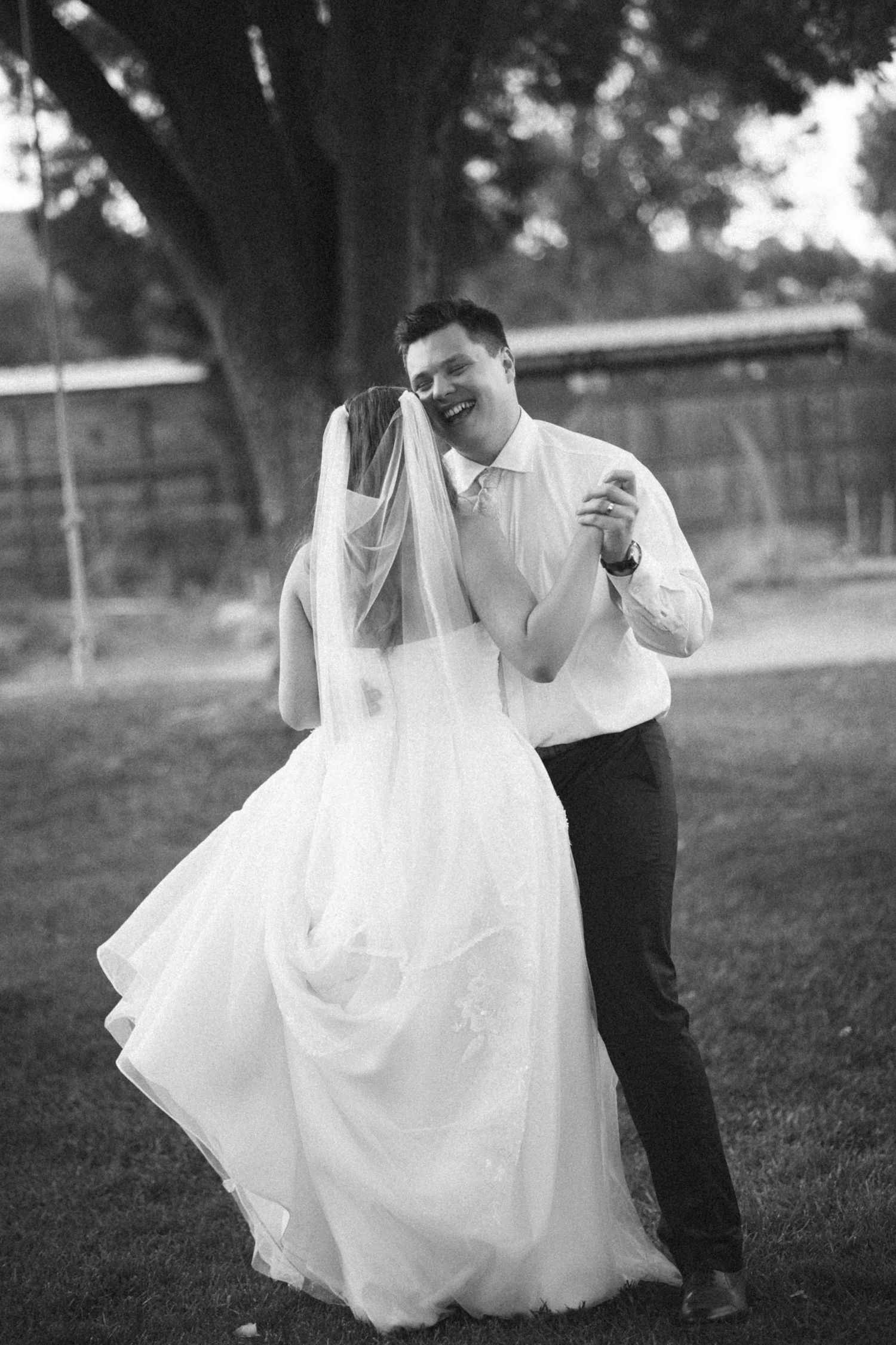 Black and white photo of a bride and groom dancing outdoors, smiling joyfully on their wedding day.