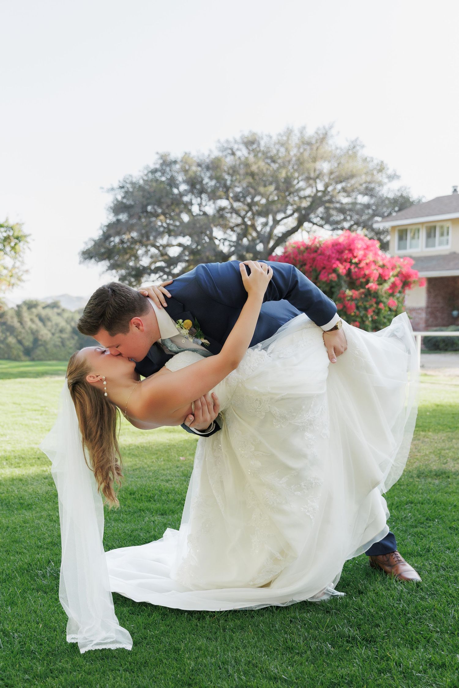 Bride lifts groom in a romantic dip kiss on green lawn, white wedding gown flowing, pink flowers and trees in background.
