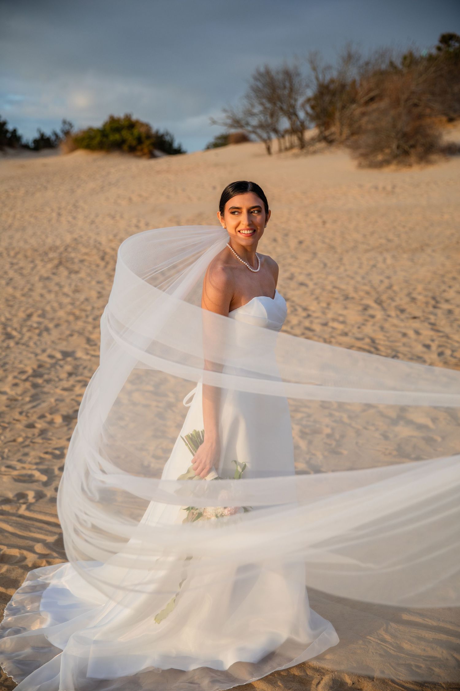 A flowing white wedding dress billows in the wind on a sandy beach during a sunset photoshoot.
