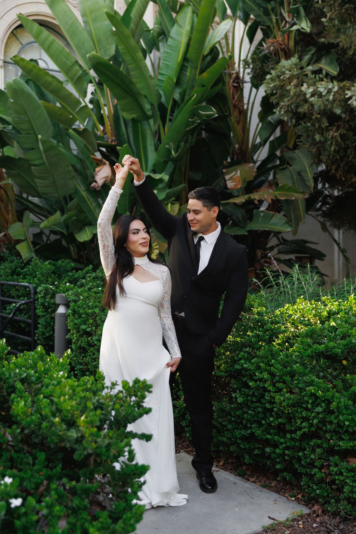 Bride in white gown and groom in black suit dancing outdoors among lush tropical greenery.