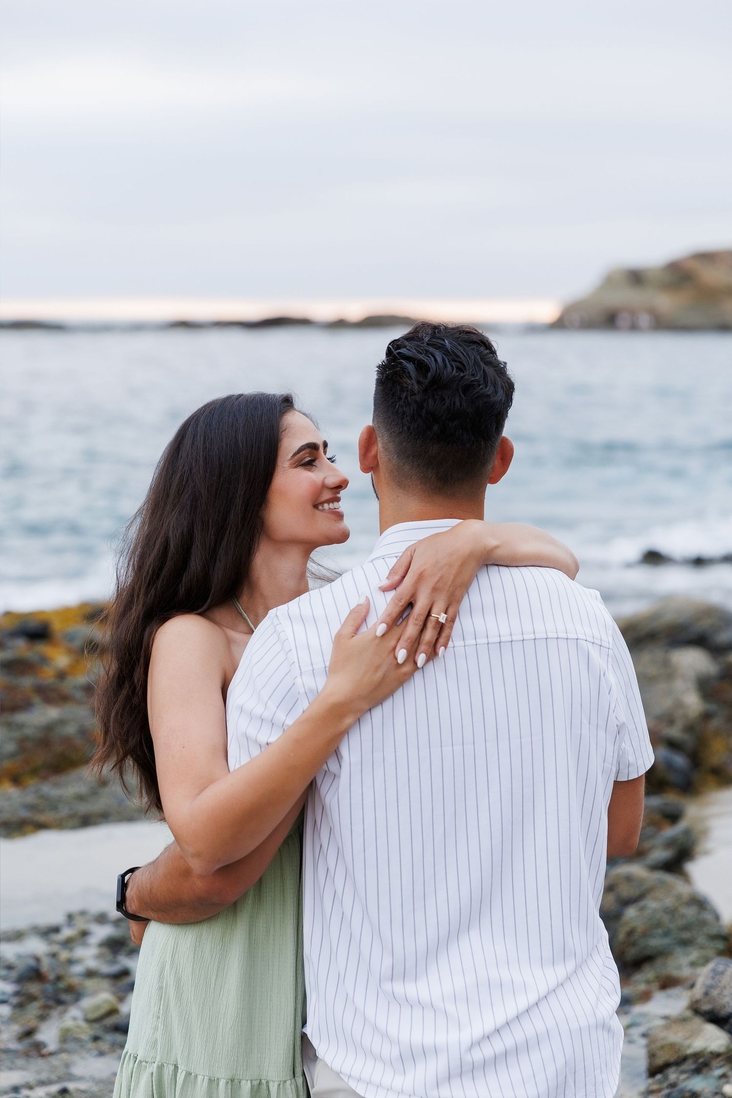 A smiling woman with dark hair embraces a man in a striped shirt on a rocky beach at dusk.
