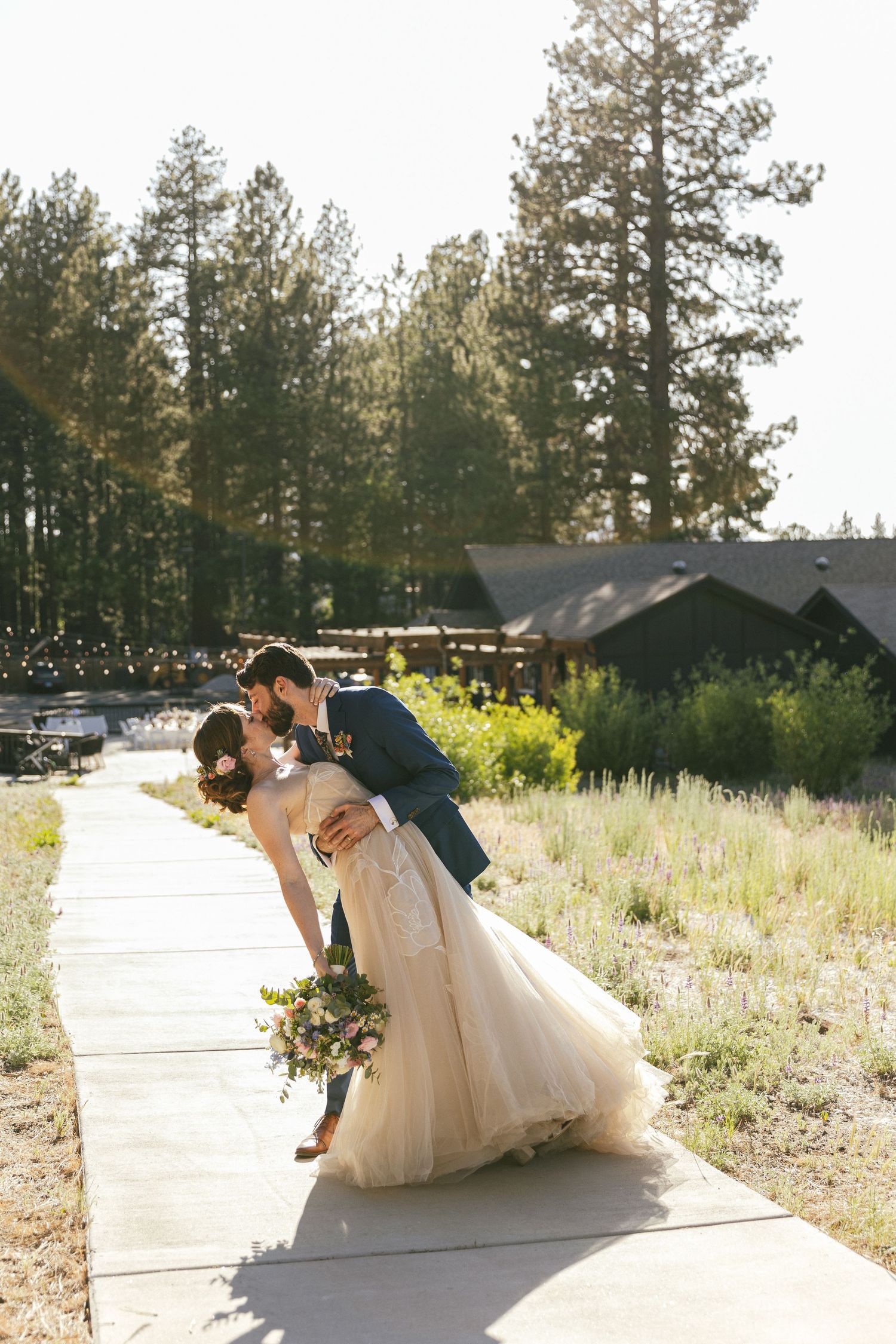 Romantic wedding couple shares playful dips and poses on a sunlit pathway surrounded by pine trees.