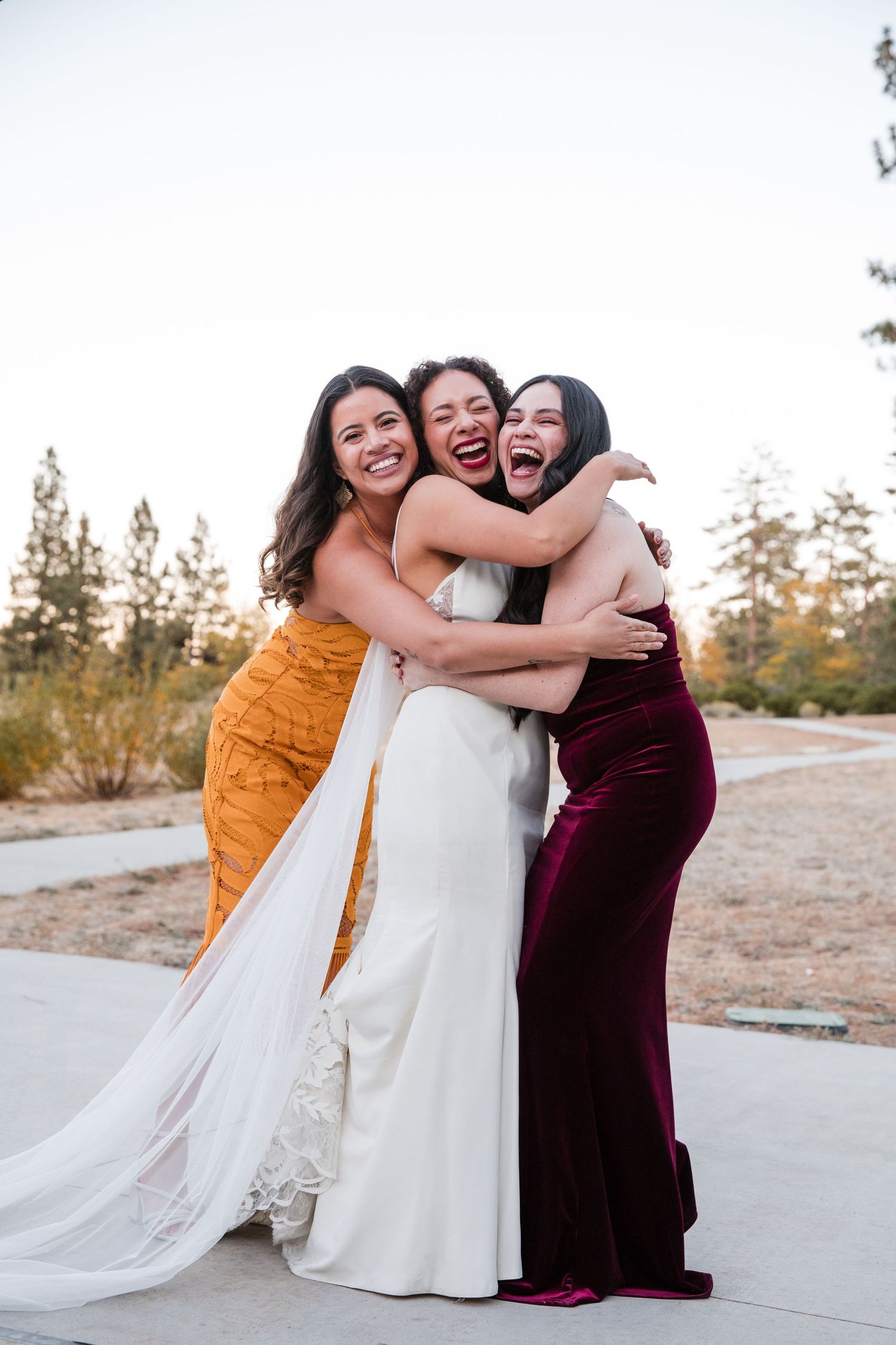 Three people in formal attire share joyful embraces and laughter during an outdoor celebration.