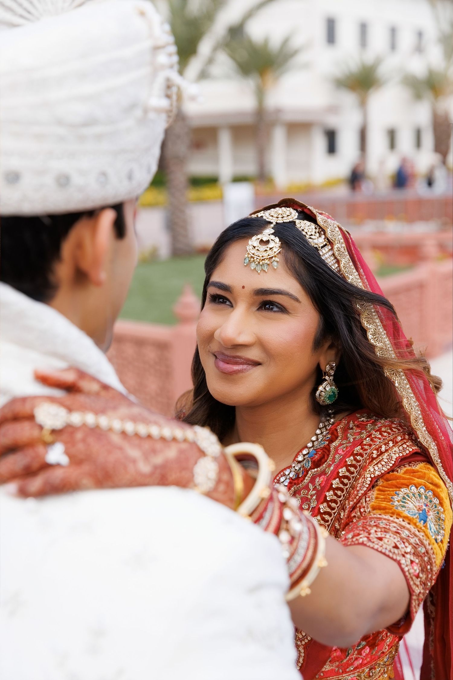 Indian bride in red saree and gold jewelry smiles at groom in white turban during traditional wedding ceremony.