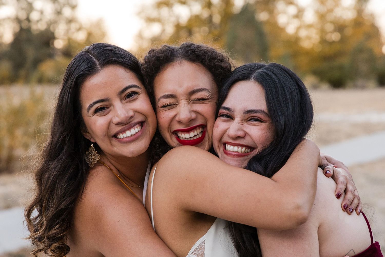 Three young women smiling and hugging each other outdoors, sharing a joyful moment together.