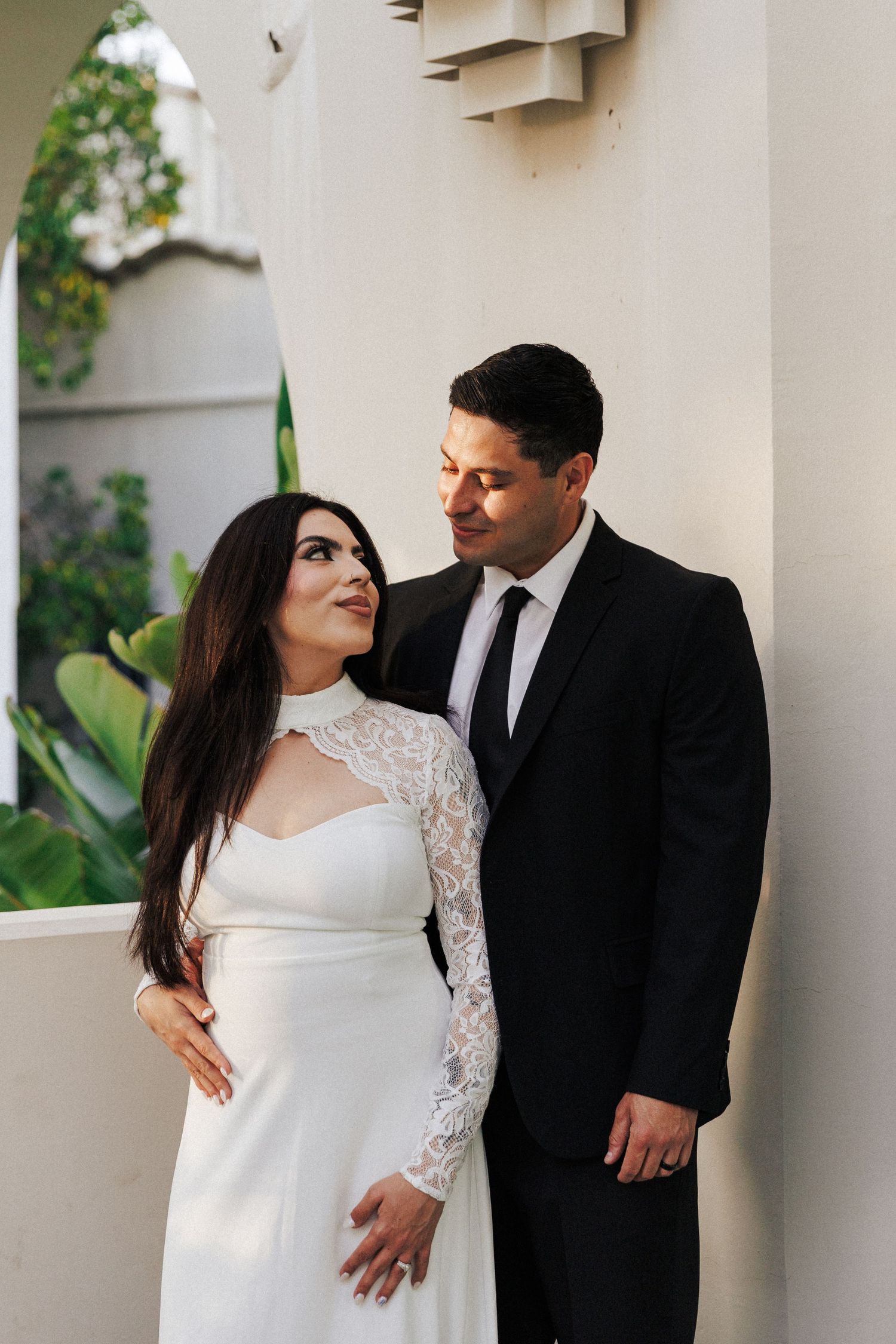 Elegant couple in wedding attire, bride in white lace gown and groom in black suit, posing outdoors.