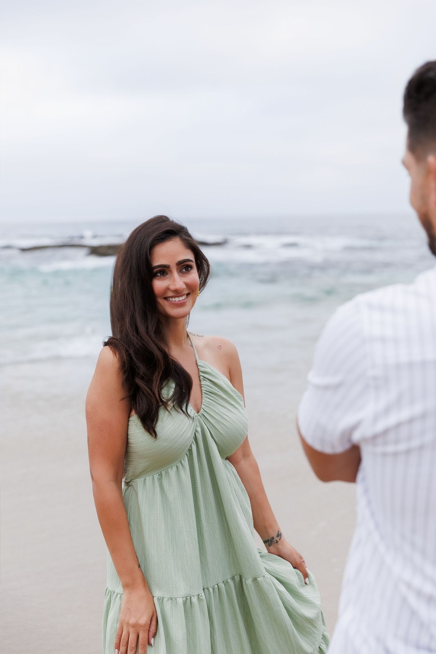 A smiling woman in a sage green halter maxi dress stands on a beach with ocean waves behind her.
