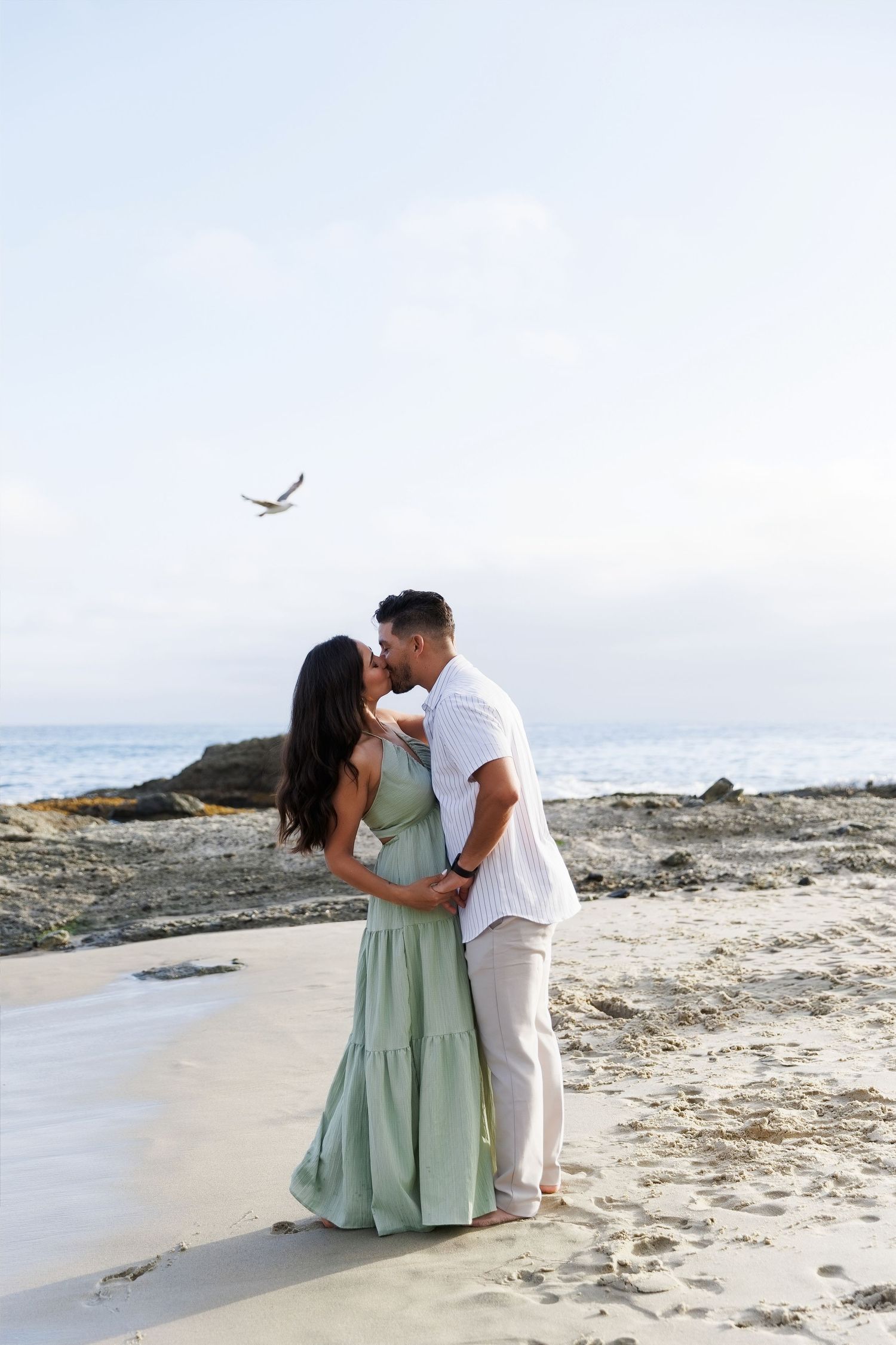 A couple shares a romantic kiss on a sandy beach, with the ocean and a seagull in the background.