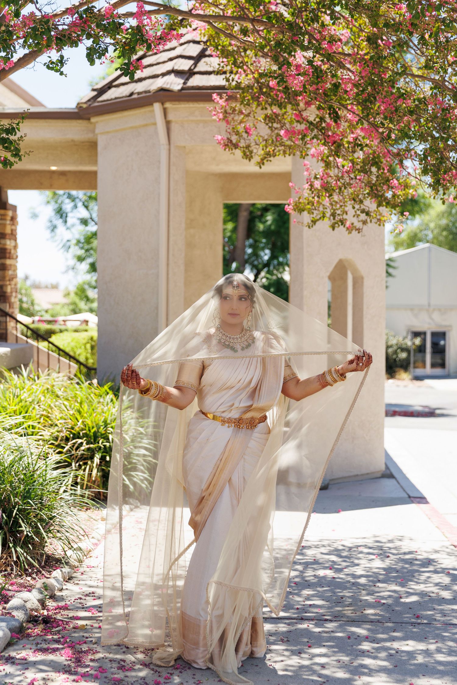 Bride in ivory saree and gold jewelry holds veil outdoors near flowering trees and beige building.