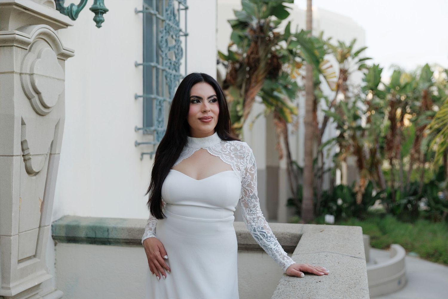 Woman in white lace long-sleeve wedding gown posing outdoors near elegant building with palm trees.