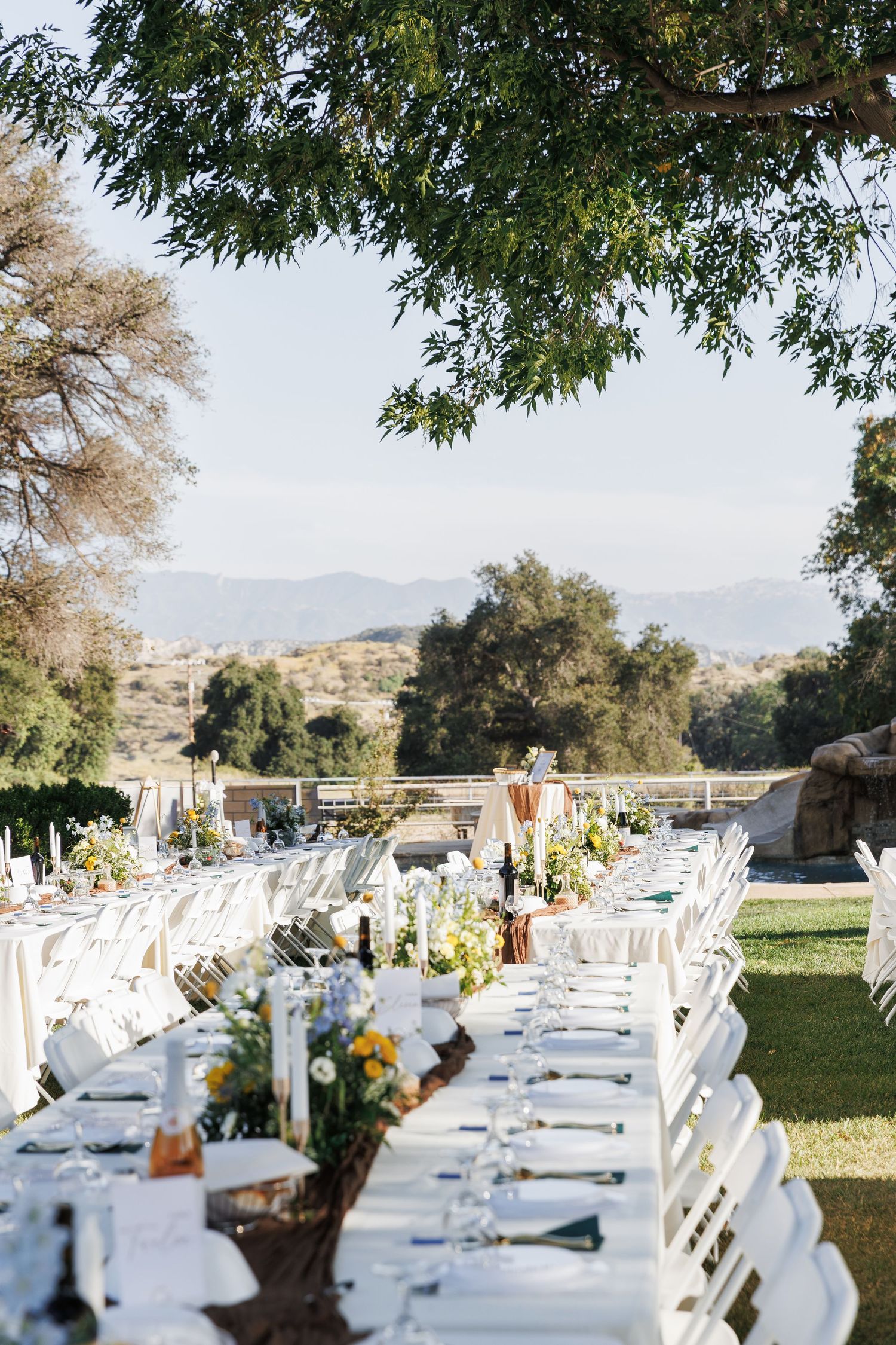 Elegant outdoor wedding reception with long white tables, floral centerpieces, and scenic mountain views under a shady tree.