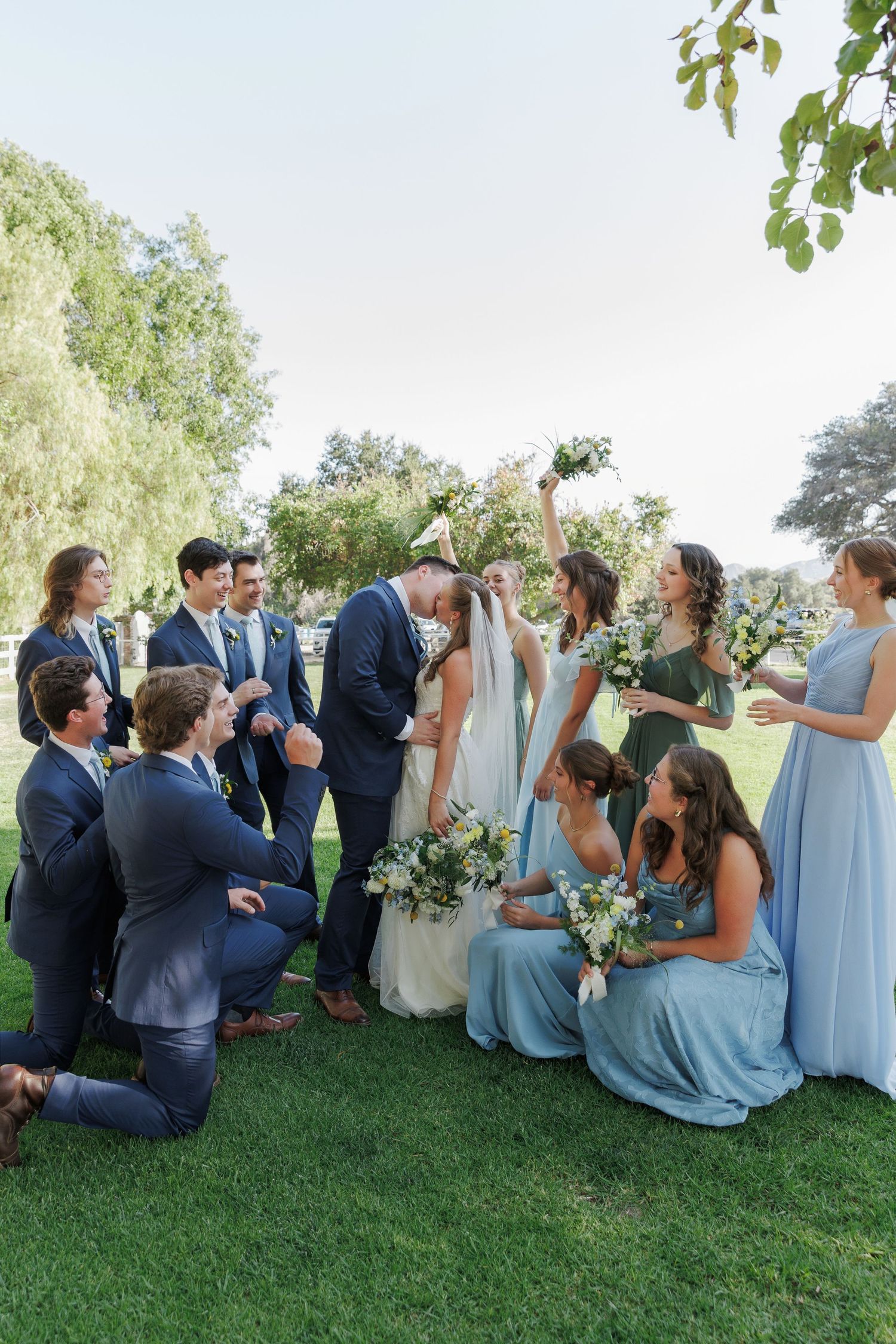 Wedding party celebrates bride and groom kissing outdoors, bridesmaids in blue dresses, groomsmen in navy suits on green lawn.