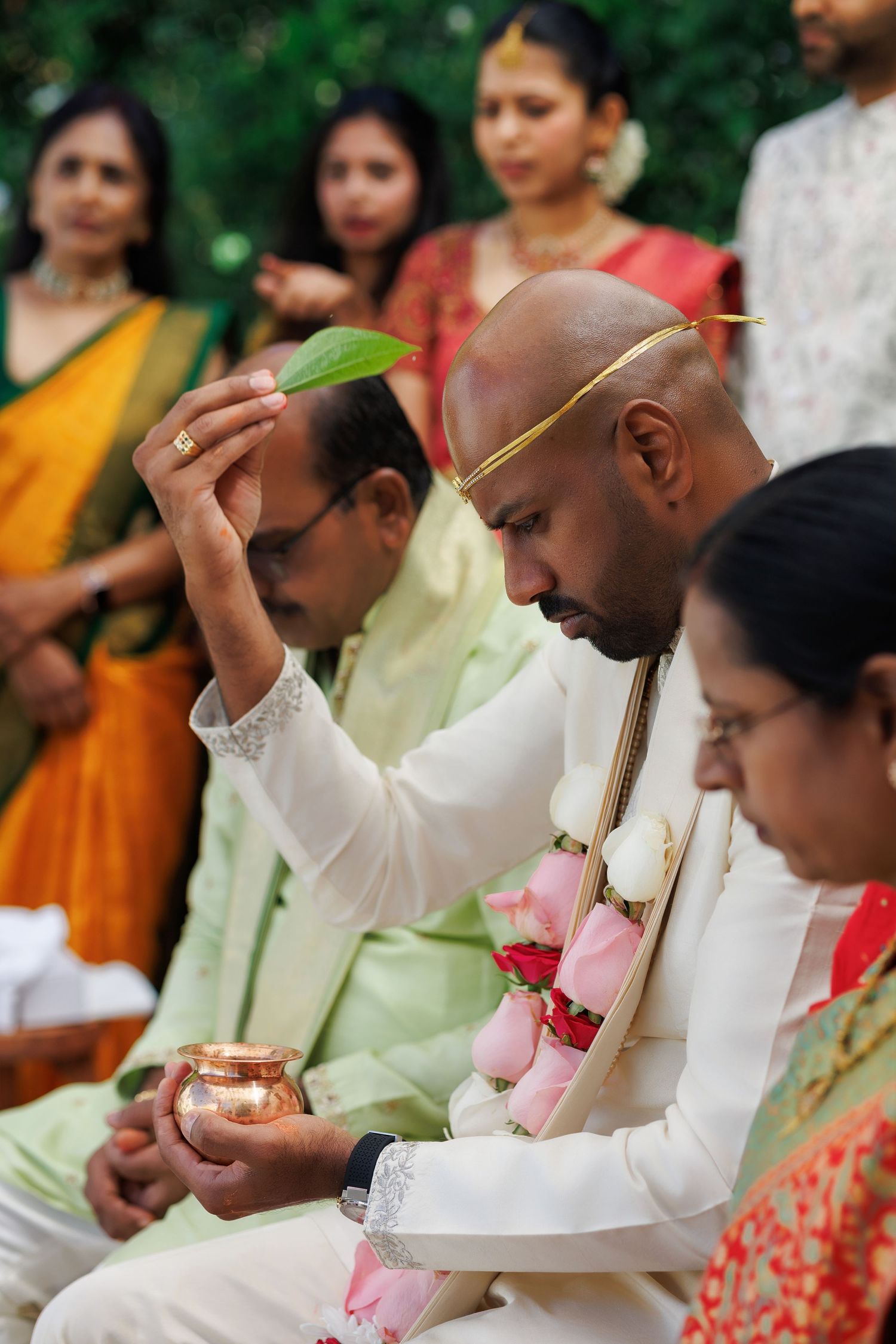 A groom in white sherwani performs Hindu wedding rituals holding a copper pot and leaf during an outdoor ceremony.
