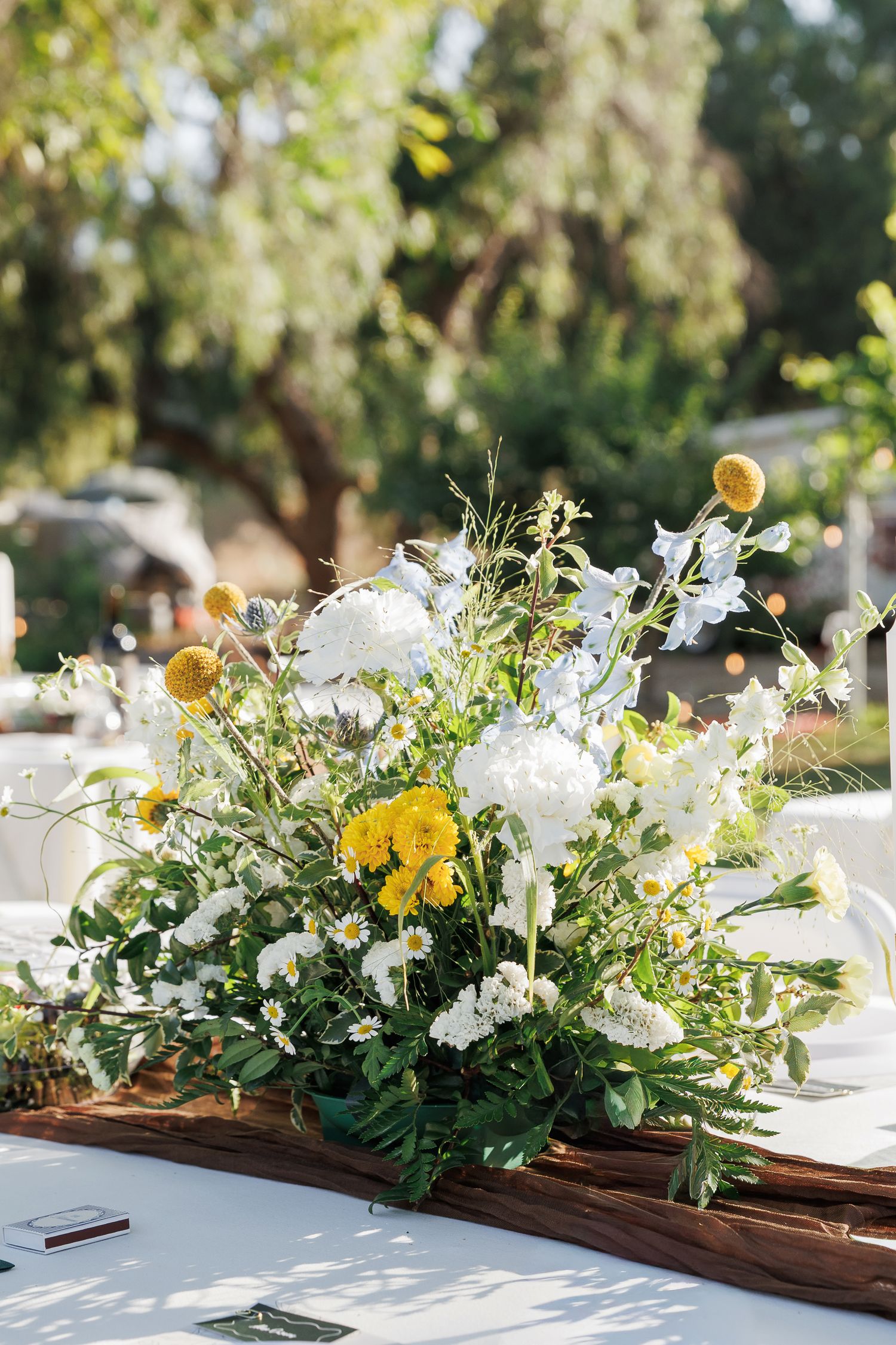 Outdoor floral centerpiece with white and yellow blooms, daisies, and greenery on a rustic wood plank table.