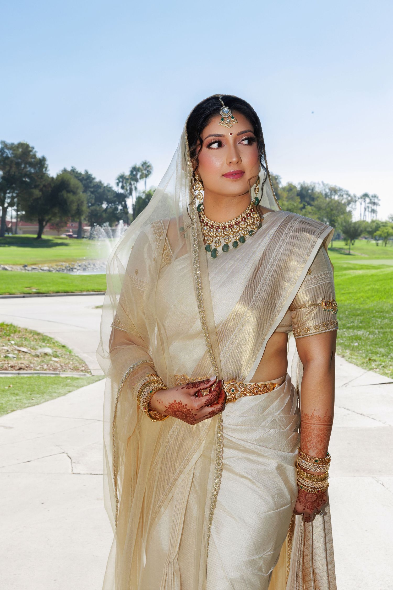 Indian bride in ivory gold saree with traditional jewelry, henna hands, and veil poses outdoors near fountain.