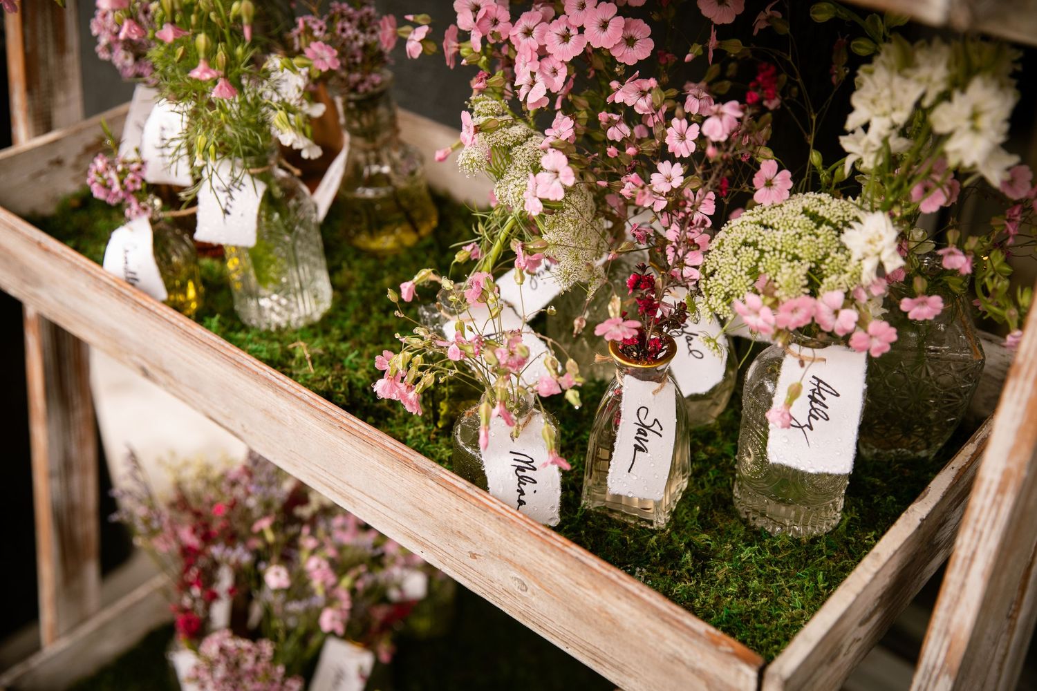 Small glass bottles filled with pink and white flowers arranged on moss-covered wooden shelves for rustic wedding decor.