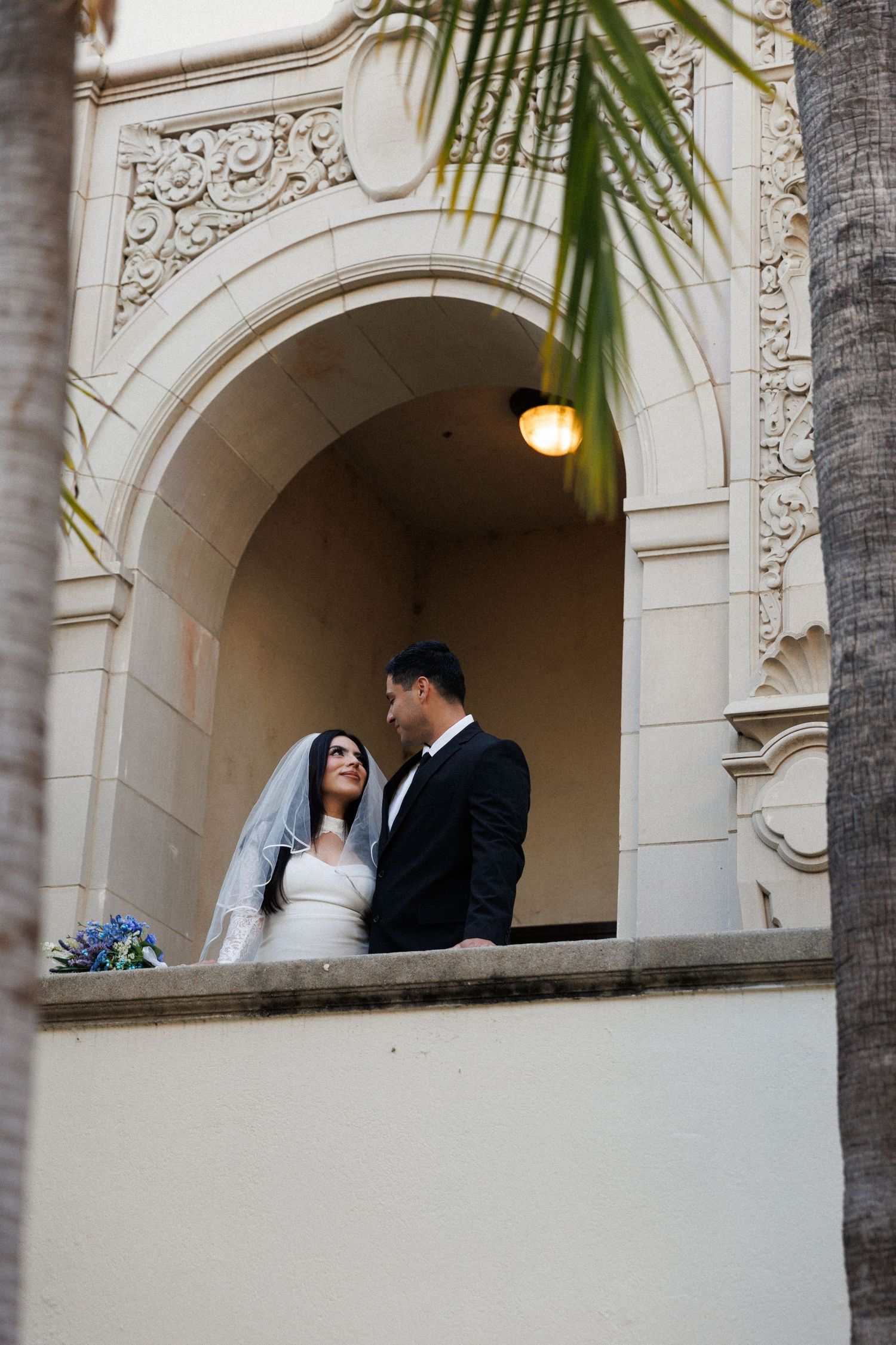 Bride and groom share a romantic moment on an ornate balcony with arched architecture at their wedding venue.