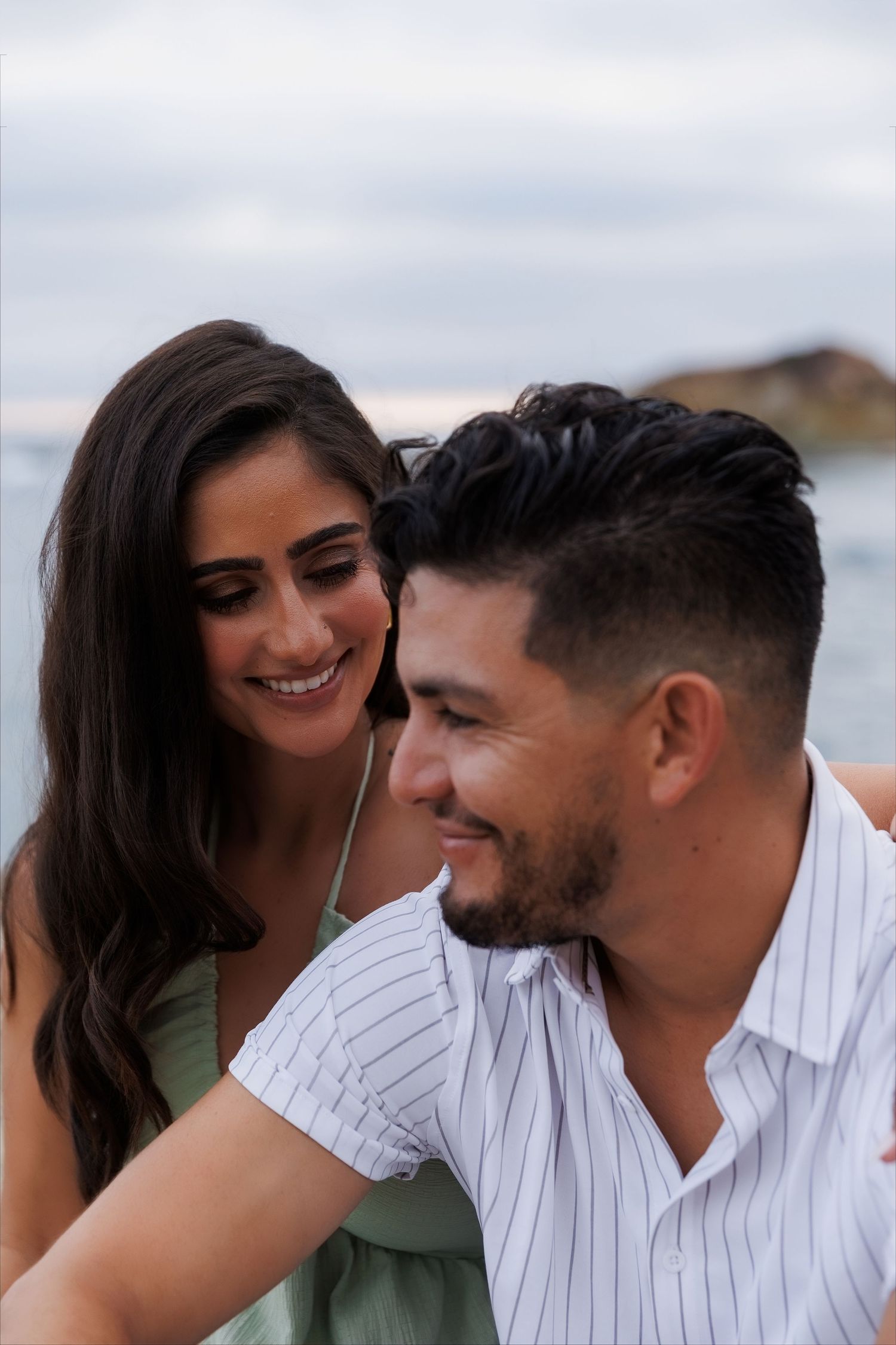 A smiling couple enjoys a romantic moment by the ocean, with the woman in a green dress leaning close to the man.