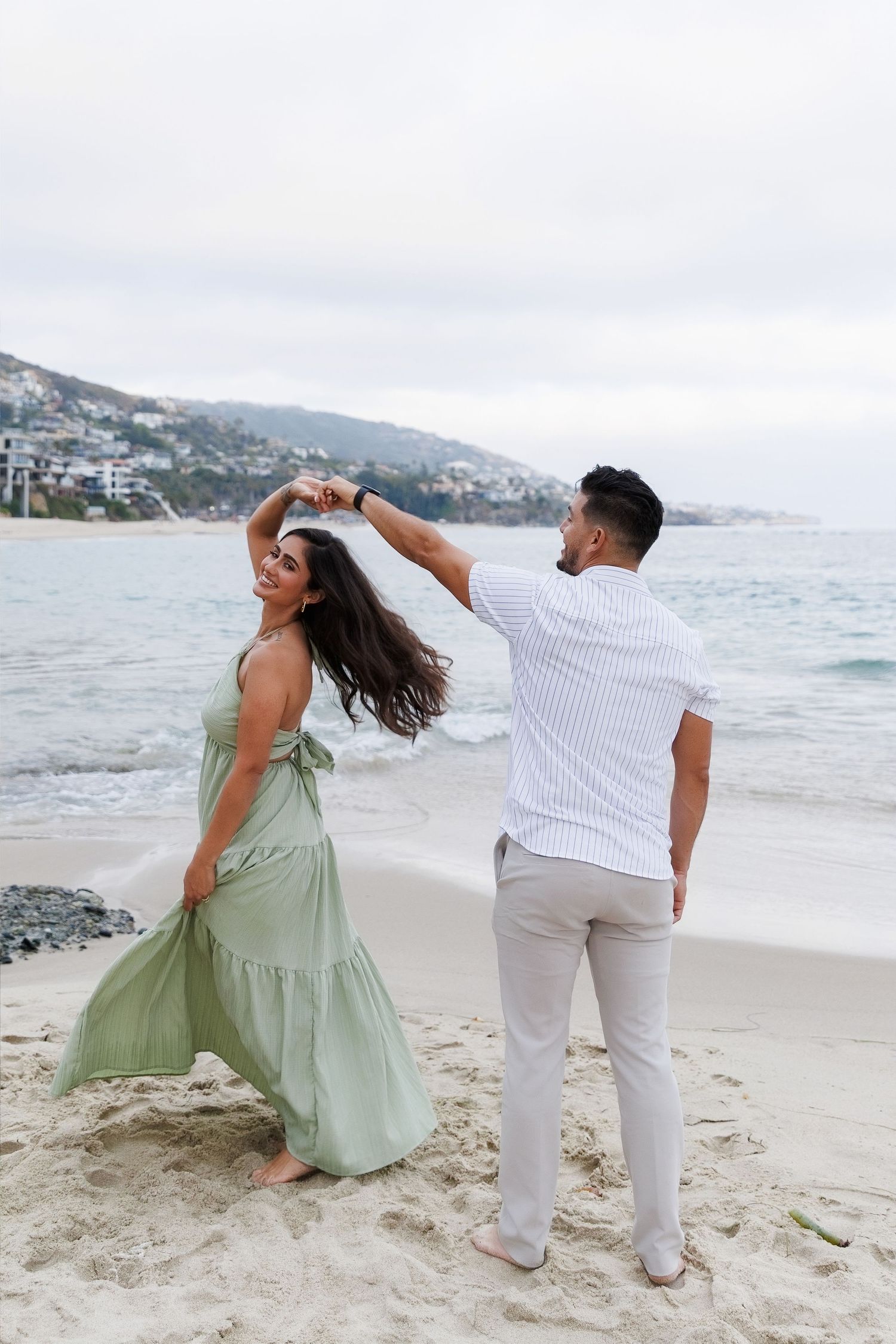 Couple dancing on a sandy beach, woman in green maxi dress being twirled by man in white striped shirt.