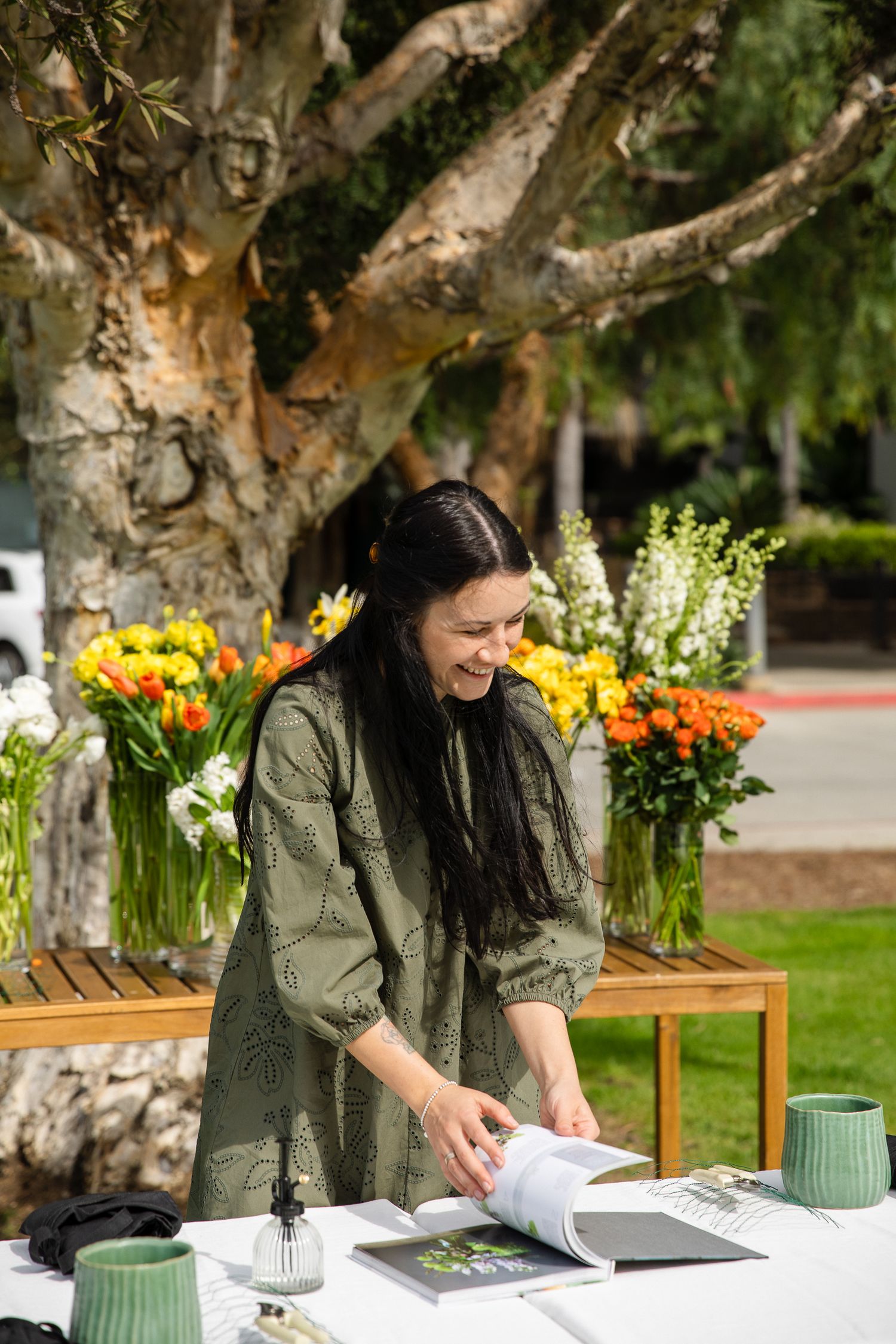 Person in olive green top working with materials at white-clothed table with floral arrangements.