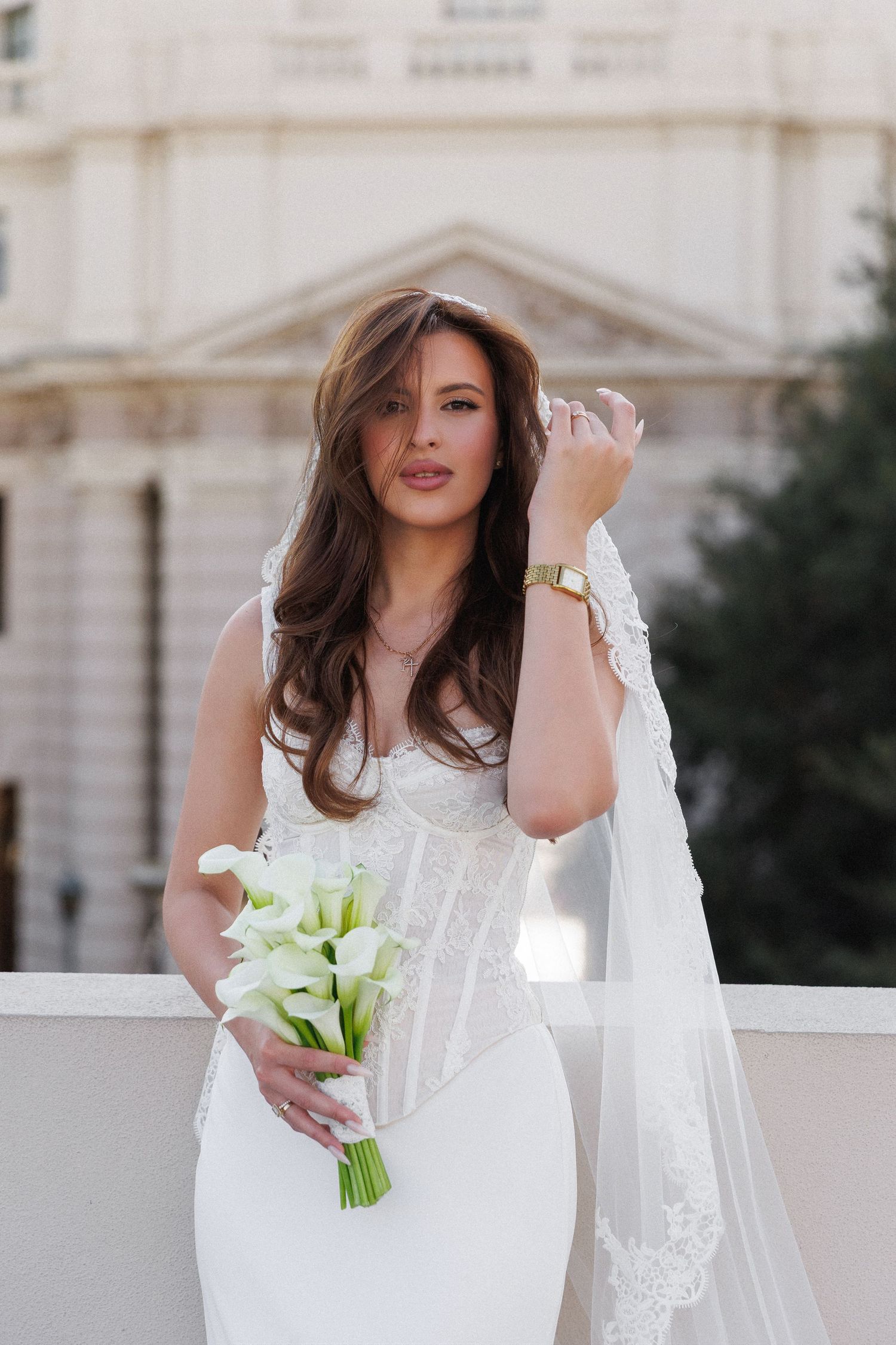 Bride in white corset gown holding calla lily bouquet with lace veil, posing outdoors near classical building.