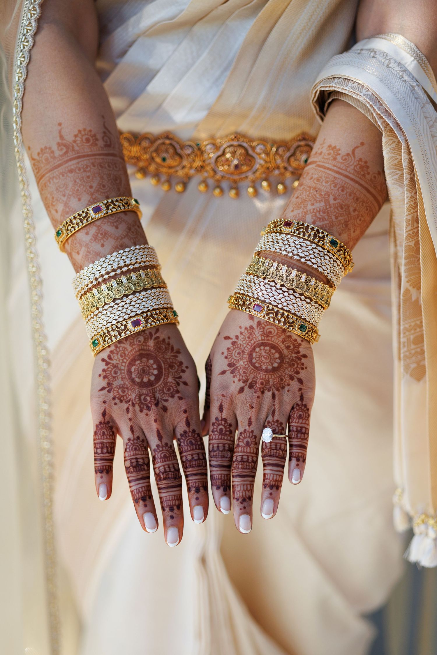 Bride showcasing intricate henna mehndi designs on hands adorned with gold bangles and rings, wearing a white saree.