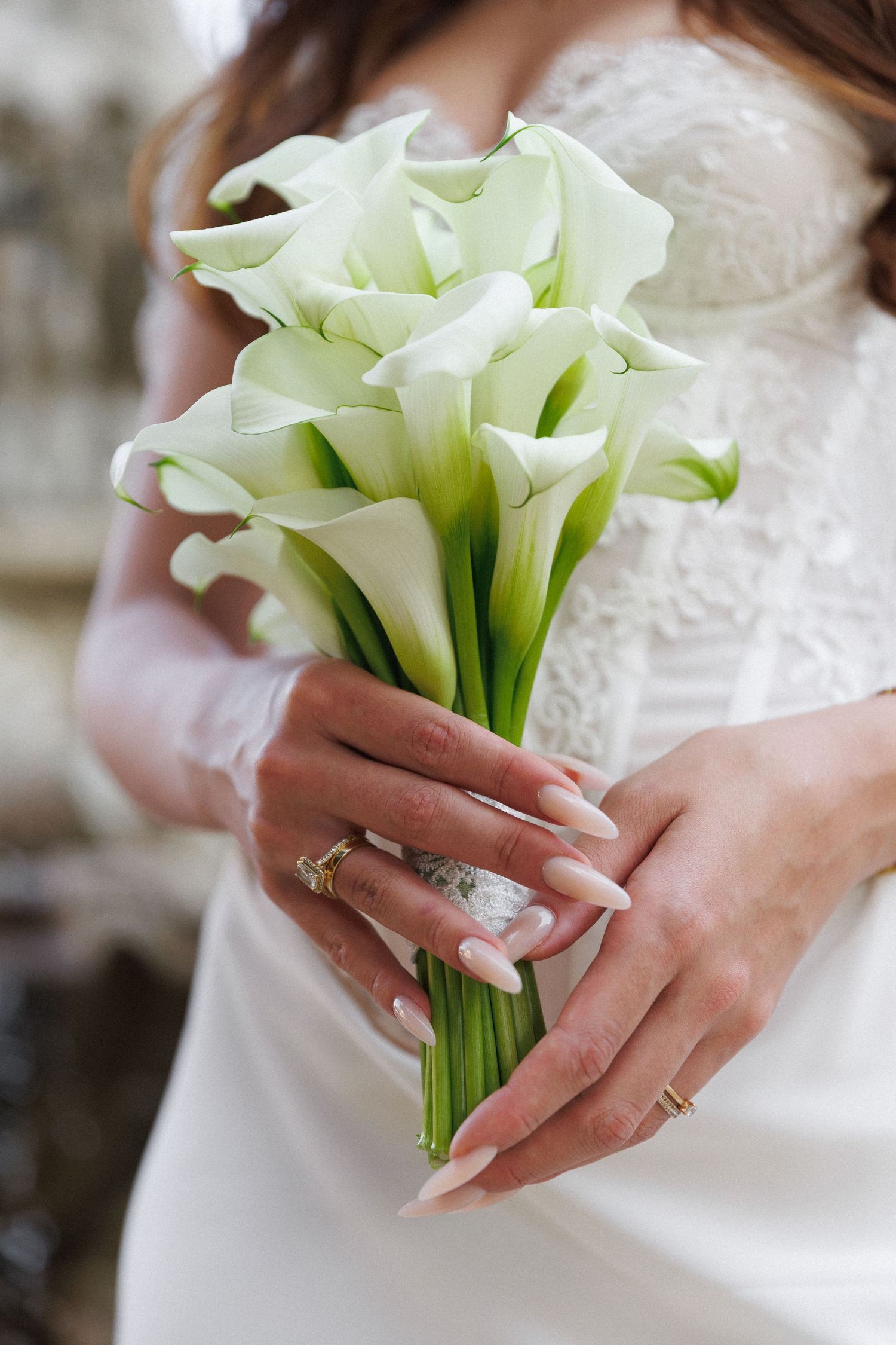 Bride holding elegant white calla lily wedding bouquet, wearing lace dress and diamond ring with manicured nails.