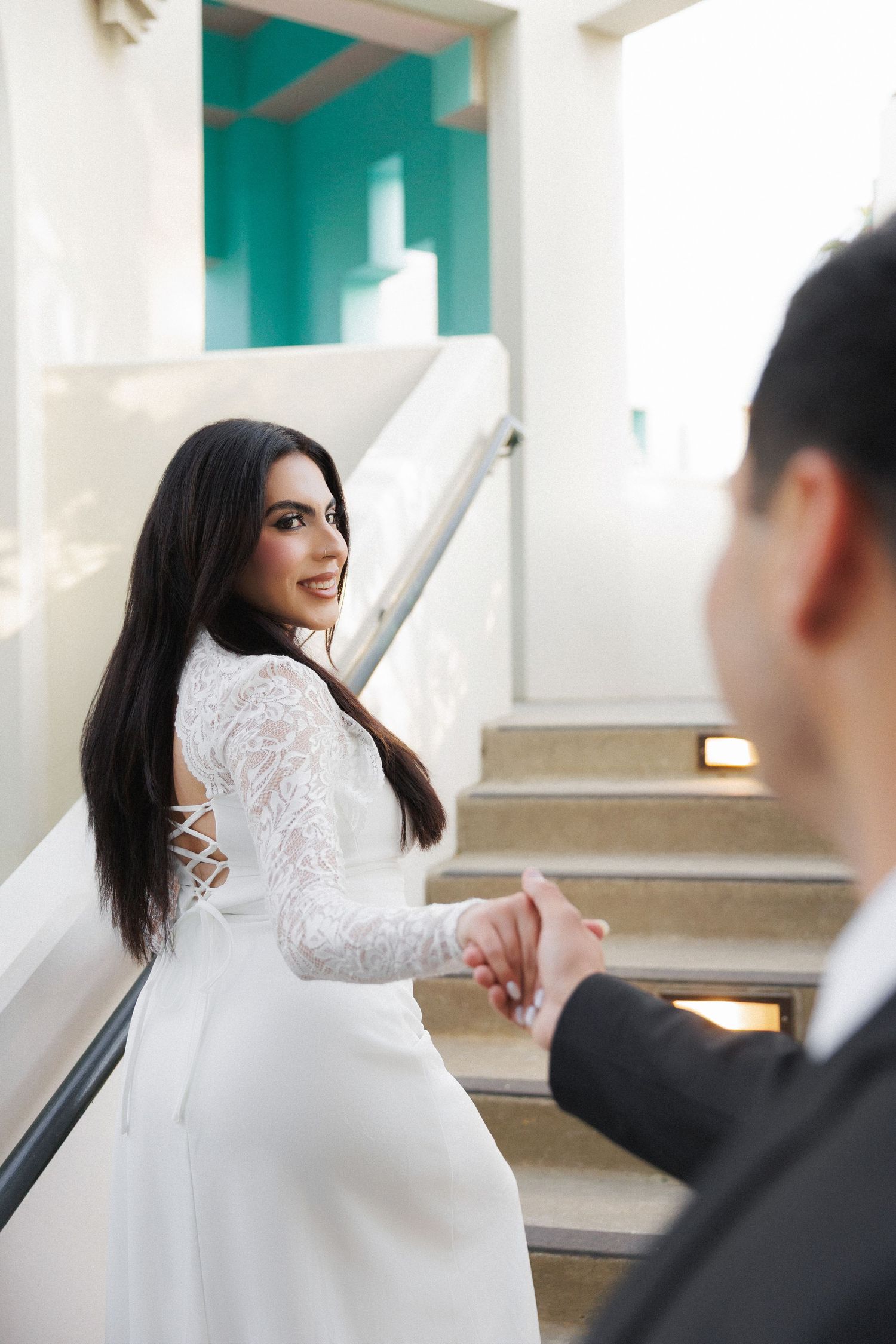 Bride in white lace gown holds groom's hand on stairs, smiling back over shoulder in romantic wedding photo.