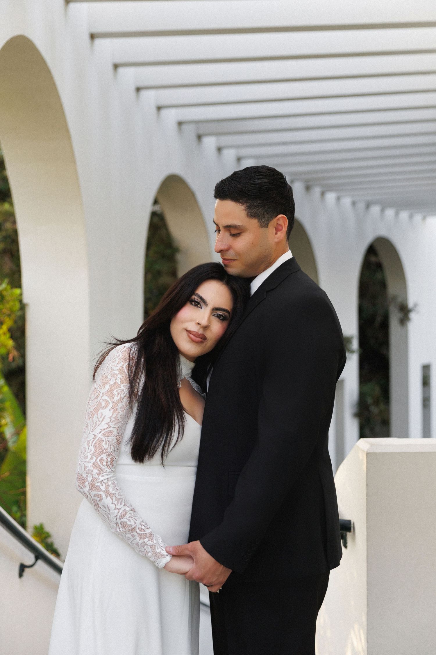 A bride in a white lace gown embraces her groom in a black suit under an elegant white arched pergola.