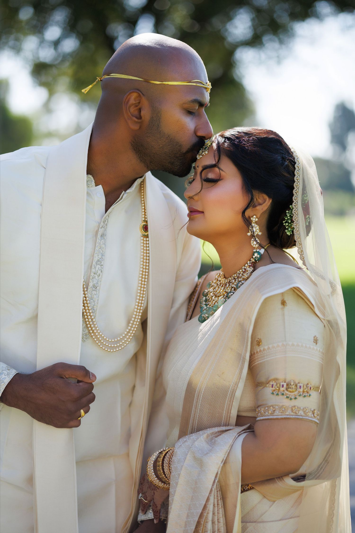 Indian bride and groom in traditional white and gold wedding attire share a tender forehead kiss outdoors.