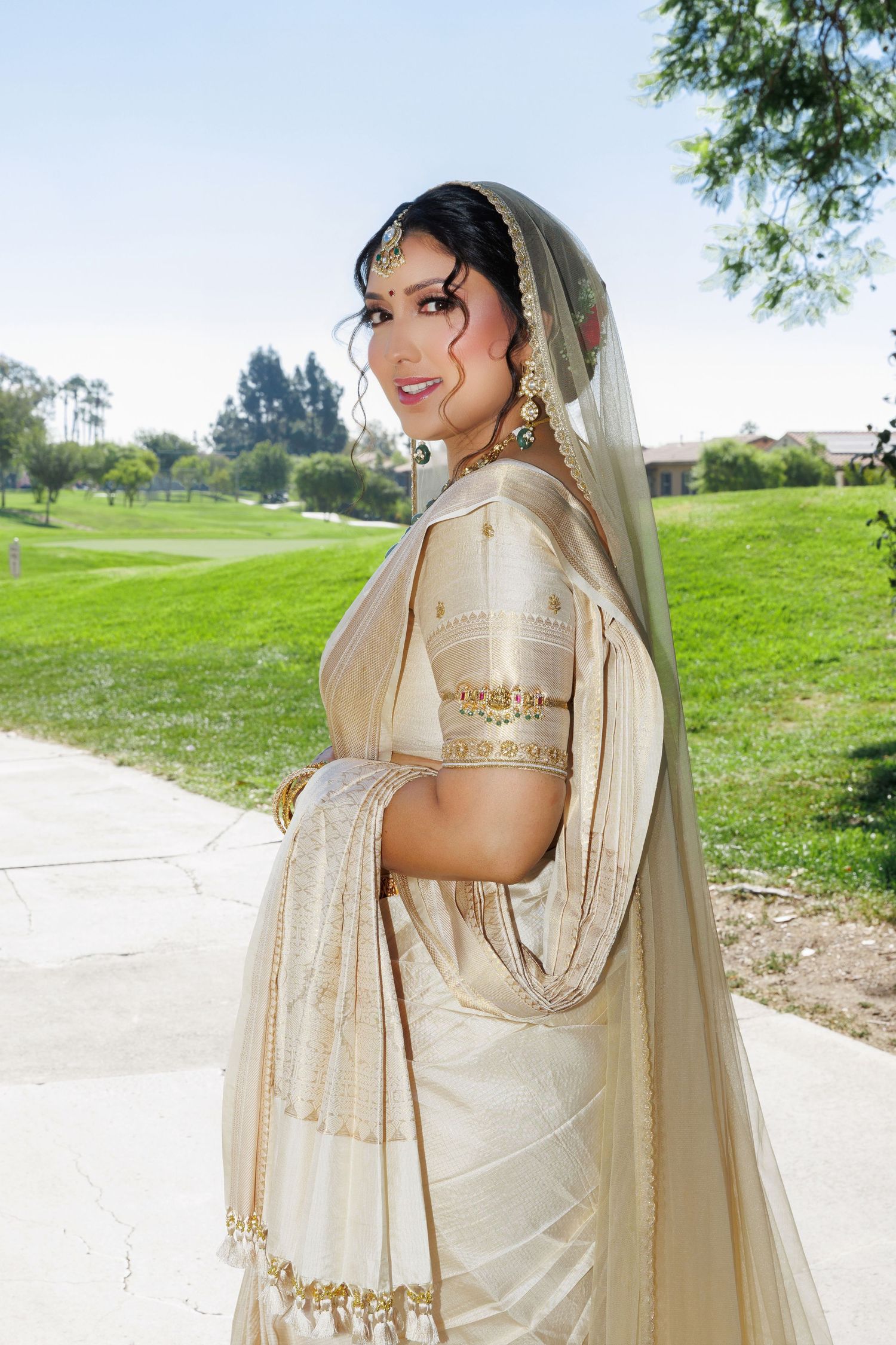 A beautiful South Asian bride in an elegant cream and gold sari with traditional jewelry and veil poses outdoors.