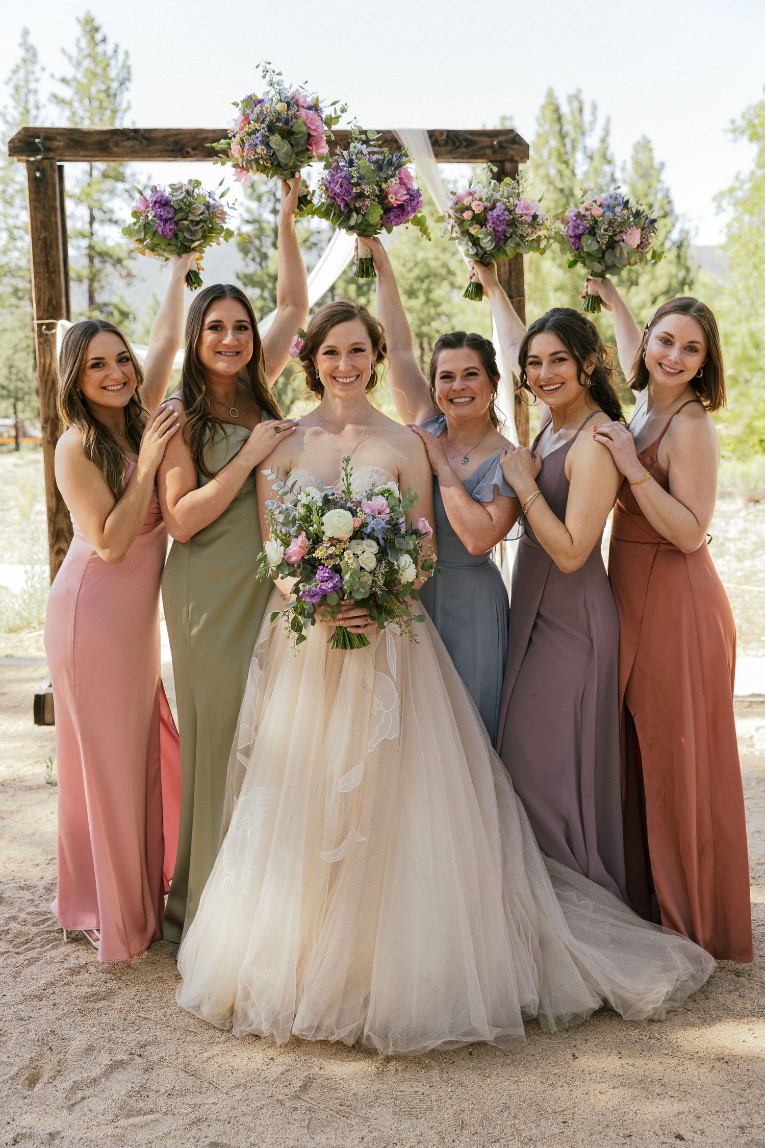 Wedding party poses outdoors wearing coordinated dresses in shades of pink, olive, and mauve with floral bouquets.
