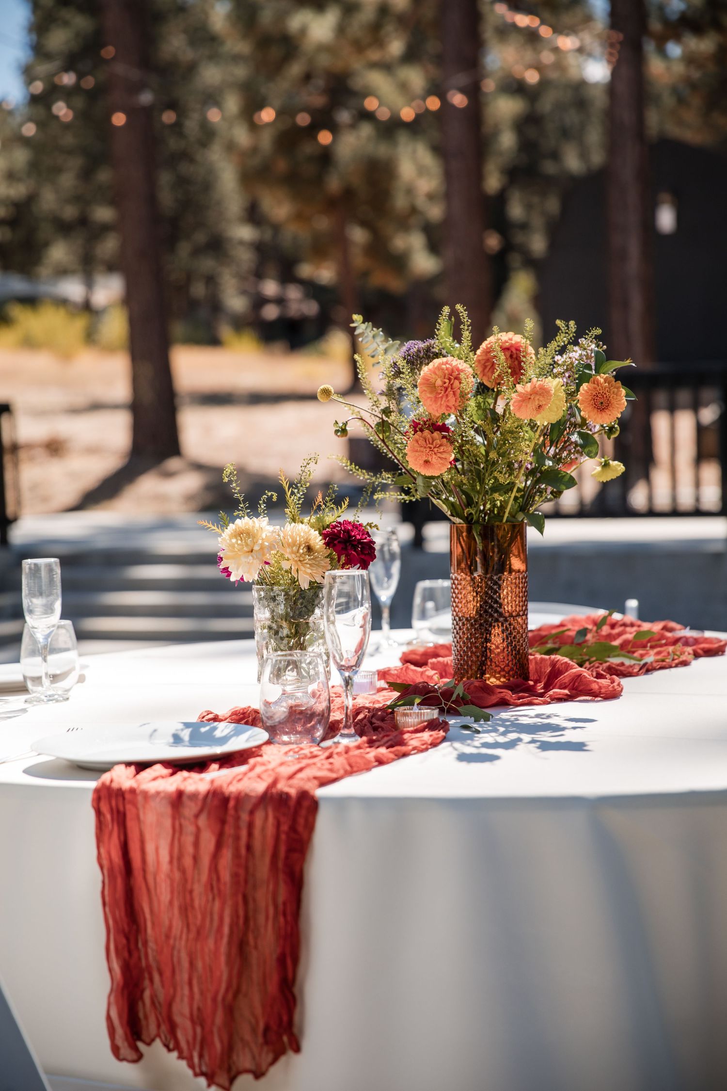 Elegant outdoor table setting with white linens, coral-colored table runners, and floral centerpieces in golden vases.