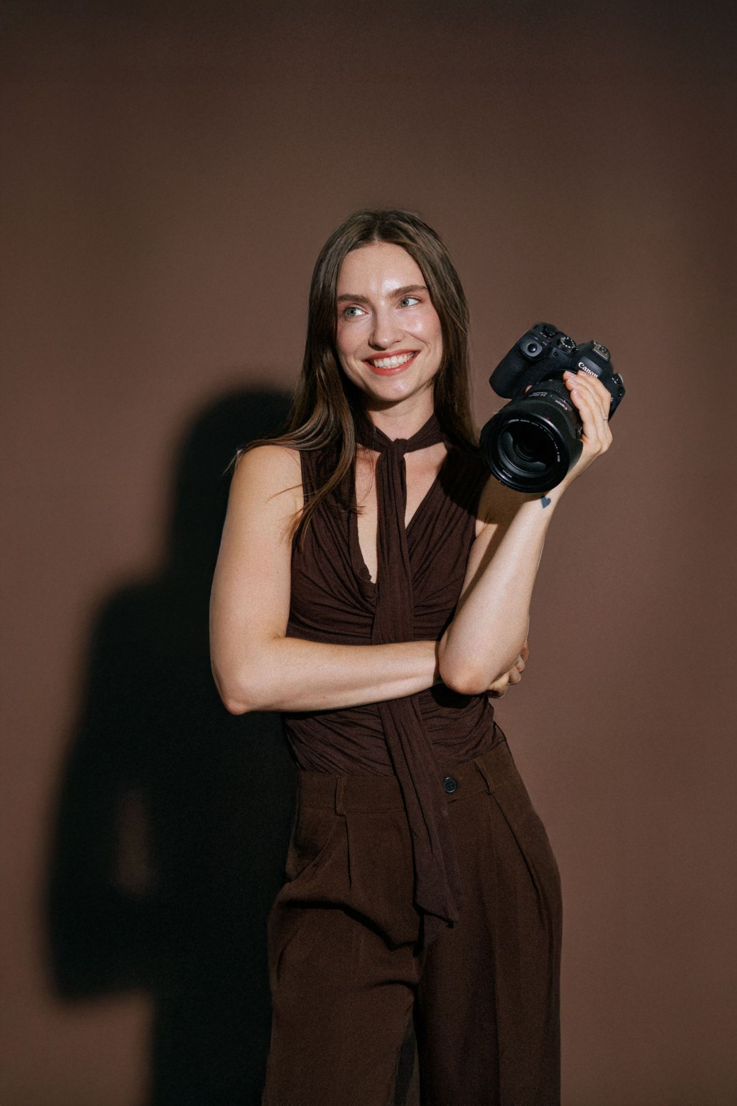 Smiling female photographer in brown outfit holding a DSLR camera against a dark brown studio background.