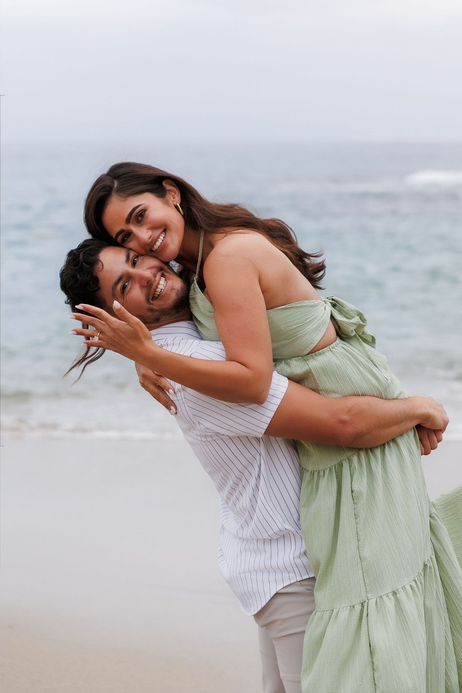 Happy couple embracing on a beach, woman in green dress showing engagement ring, smiling joyfully by the ocean.