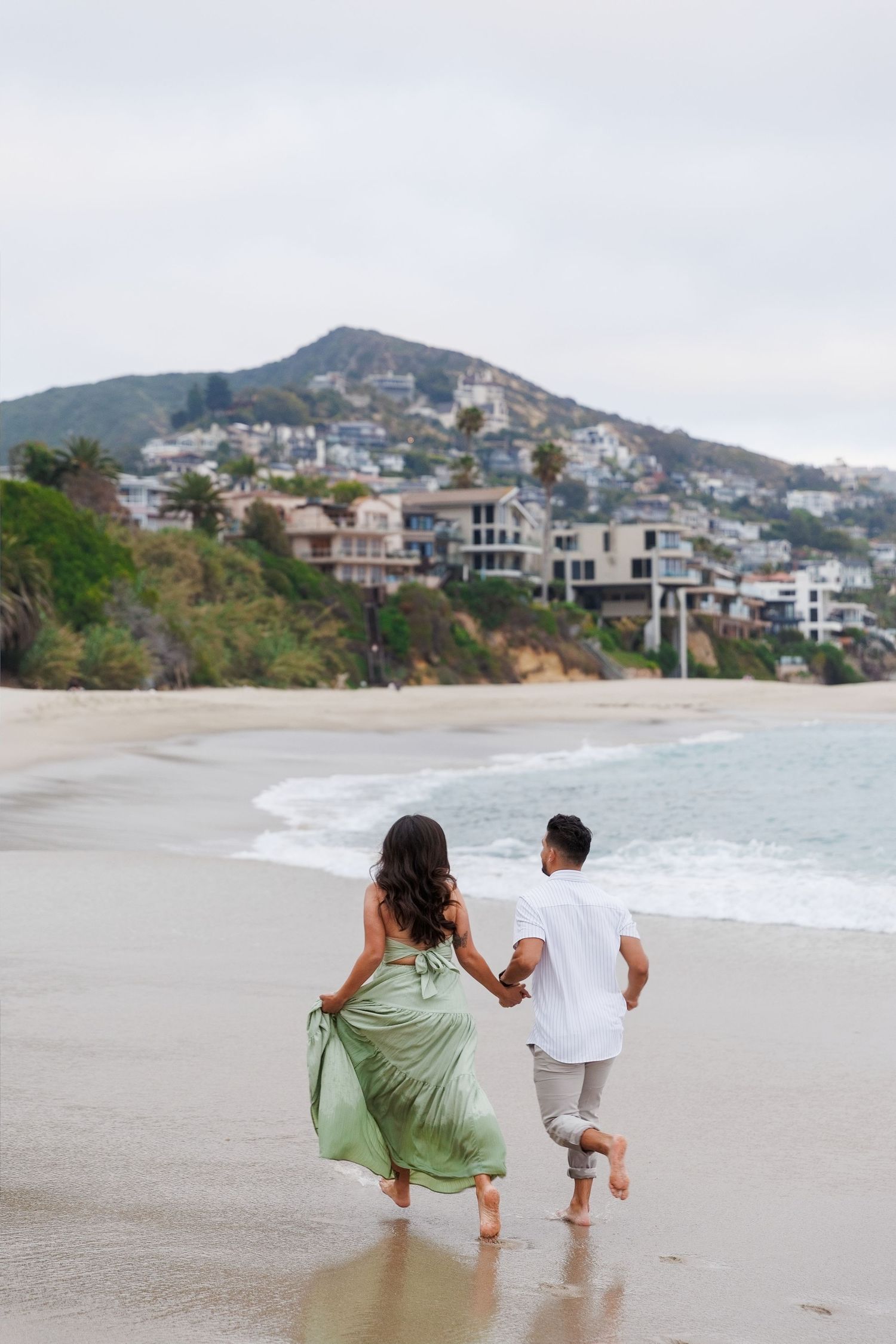 Couple holding hands running on a sandy beach with ocean waves, green hillside homes, and cloudy skies in the background.