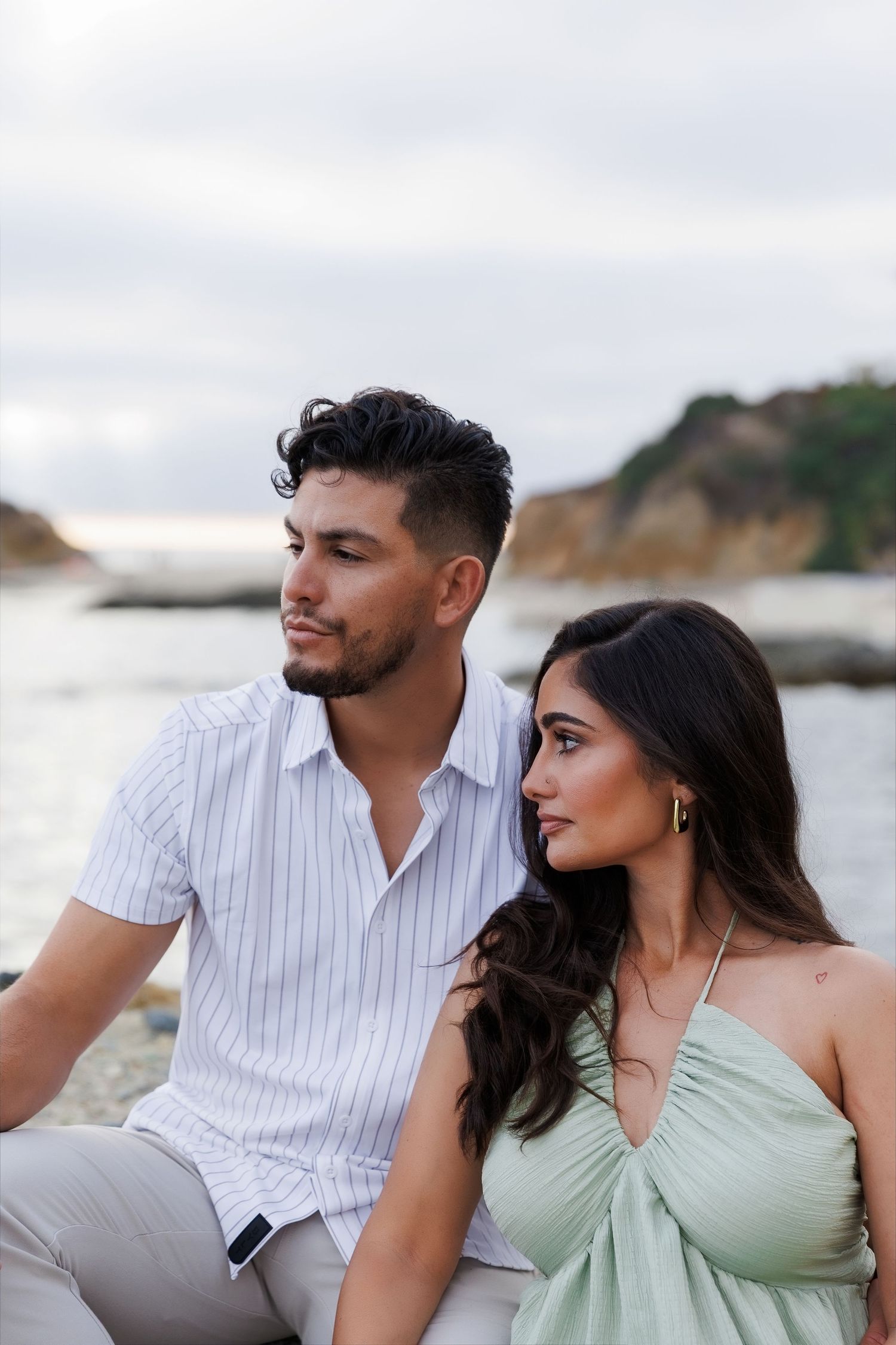 A couple sits together by a rocky coastline, the man in a striped shirt and woman in a sage green dress.