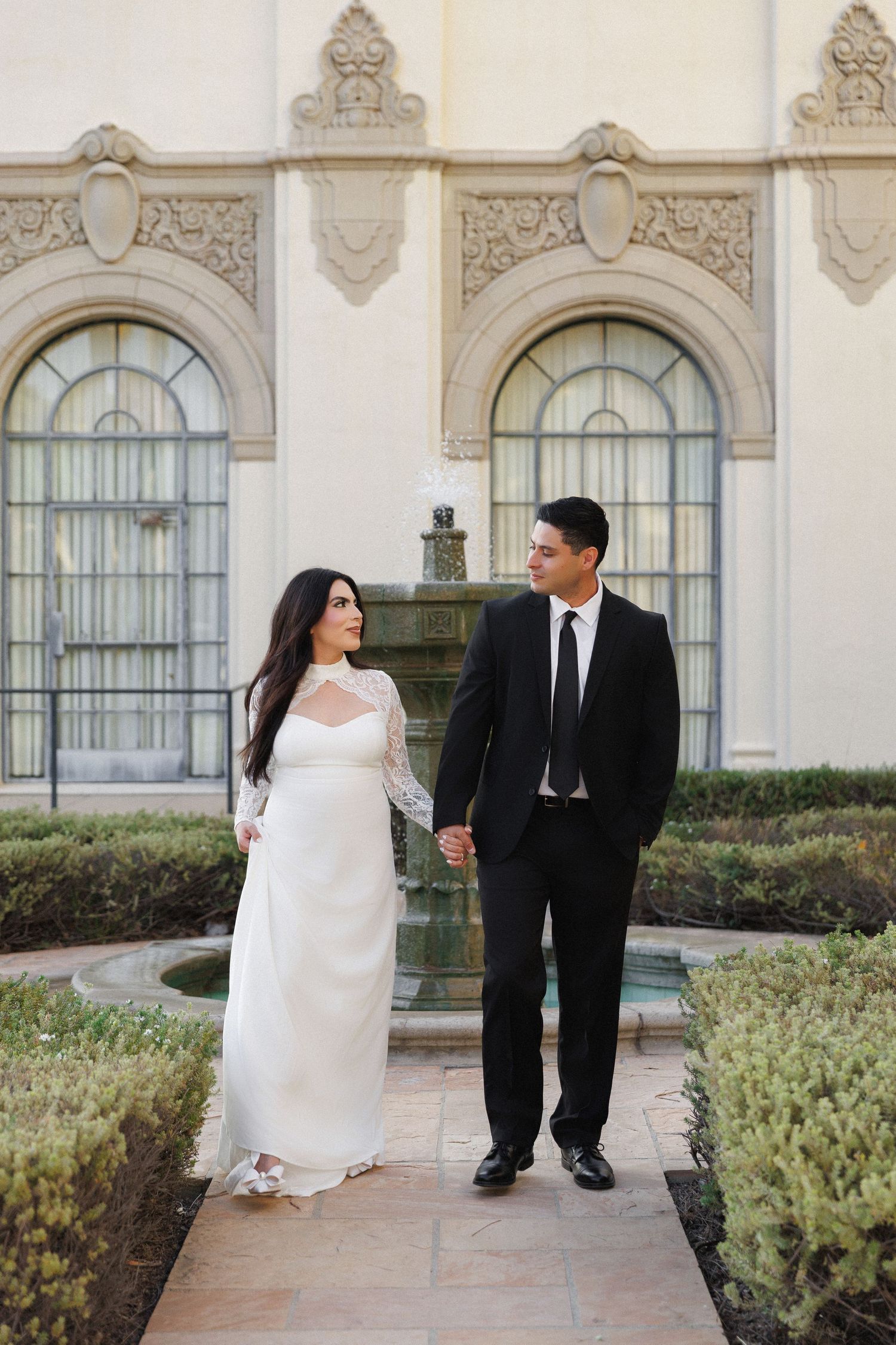 Bride in white lace gown and groom in black suit hold hands in ornate garden courtyard.