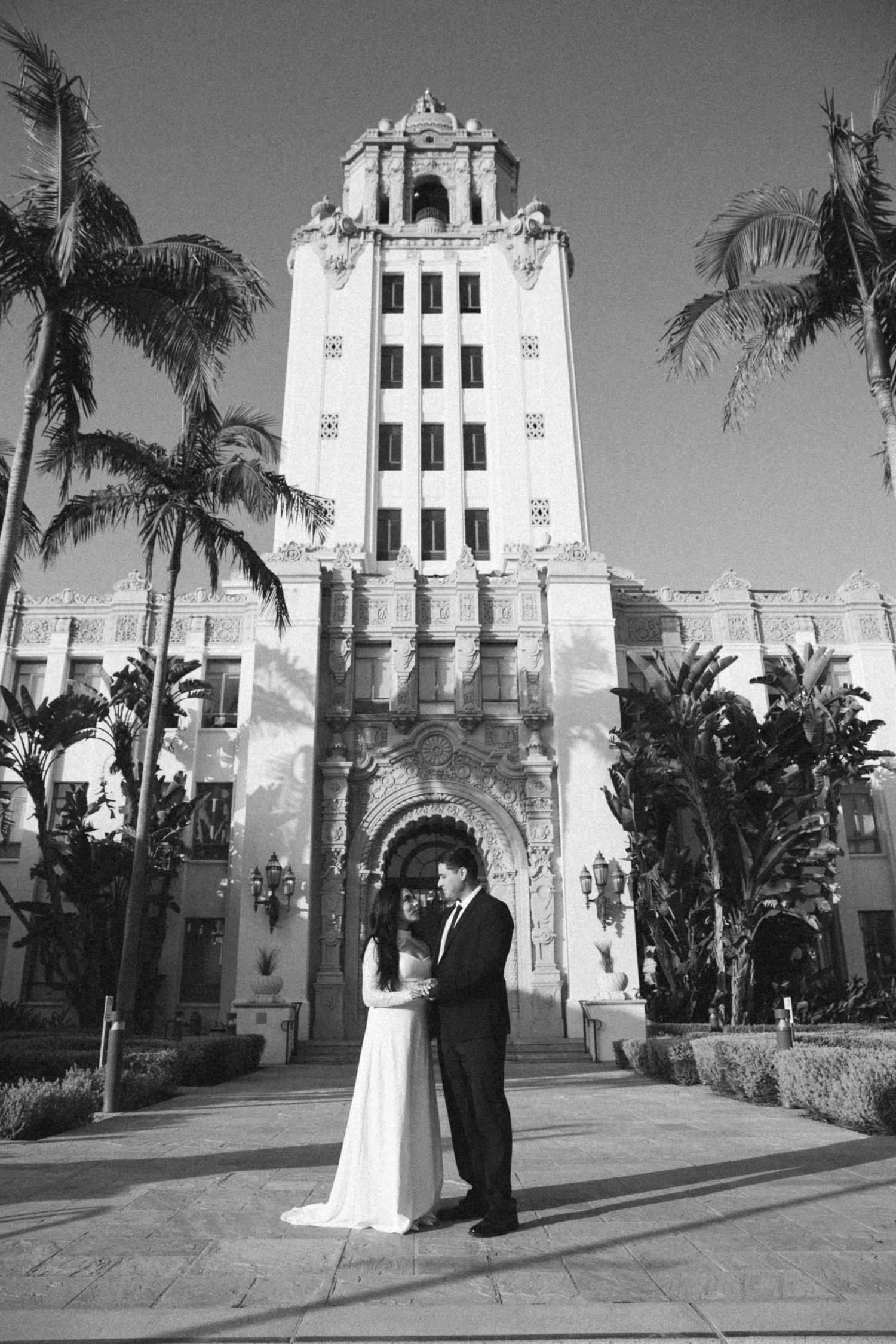 Black and white photo of a couple in wedding attire embracing in front of Beverly Hills City Hall with palm trees.
