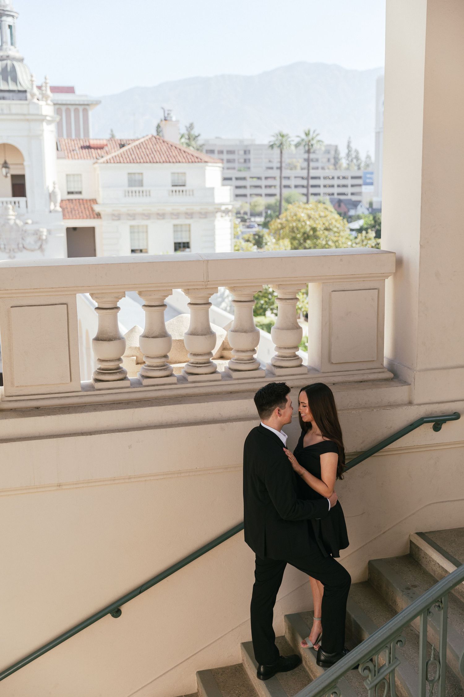 A couple sharing a tender moment on ornate white stairs with Spanish architecture in the background.