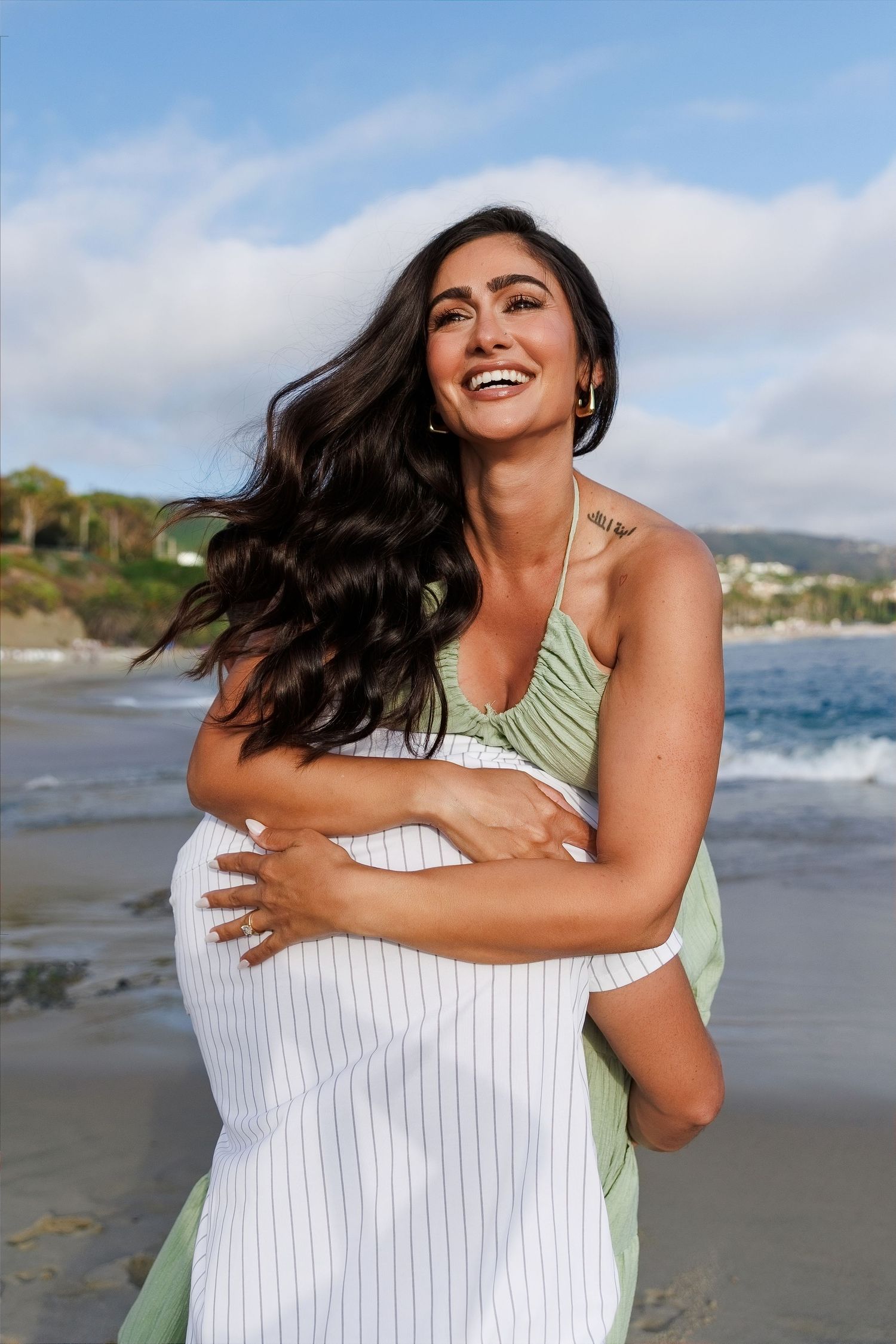 Smiling pregnant woman in green top and white skirt hugging her baby bump on a sunny beach.