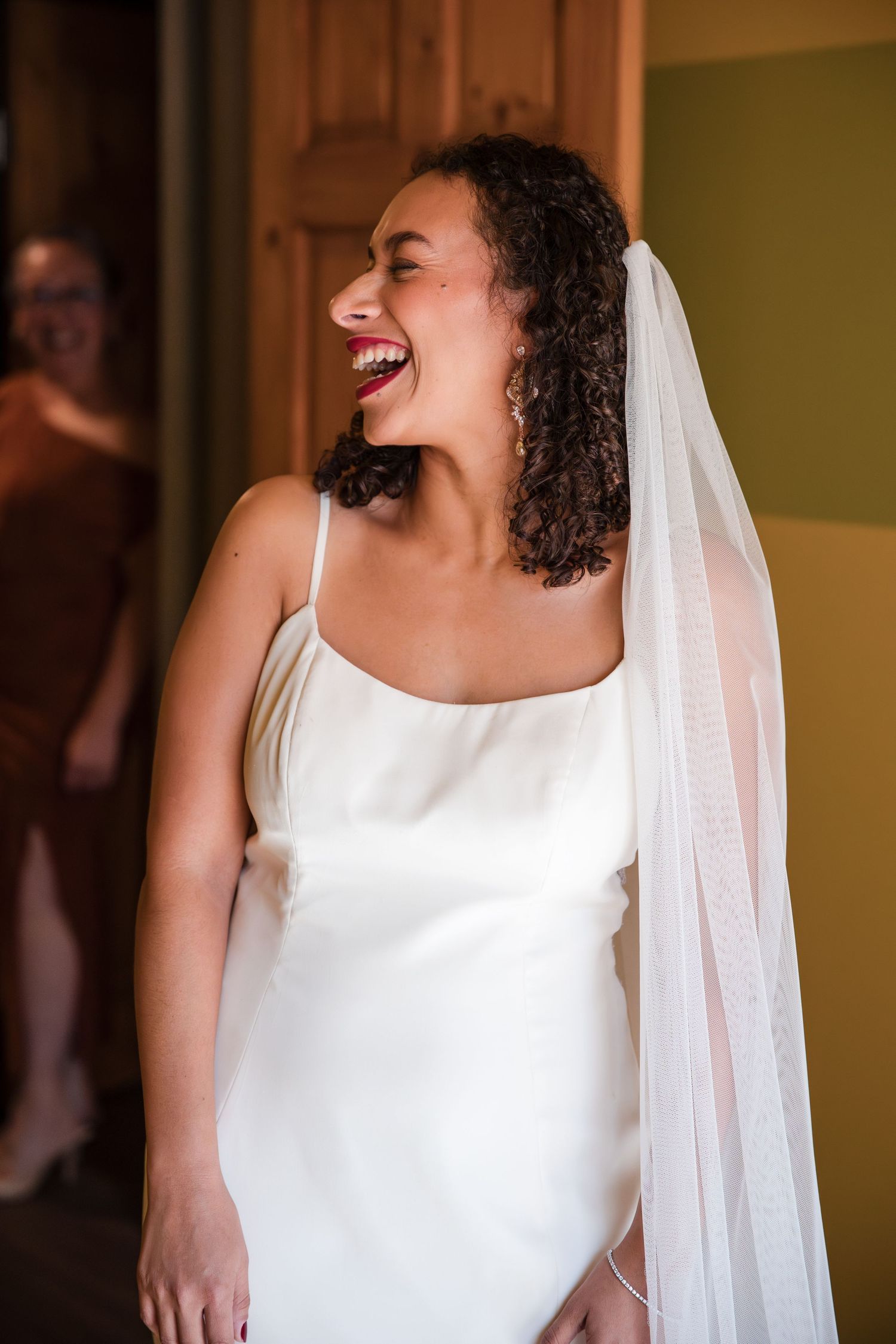 A joyful bride wearing a sleek white sleeveless wedding dress and veil laughs candidly in a warm-lit room.