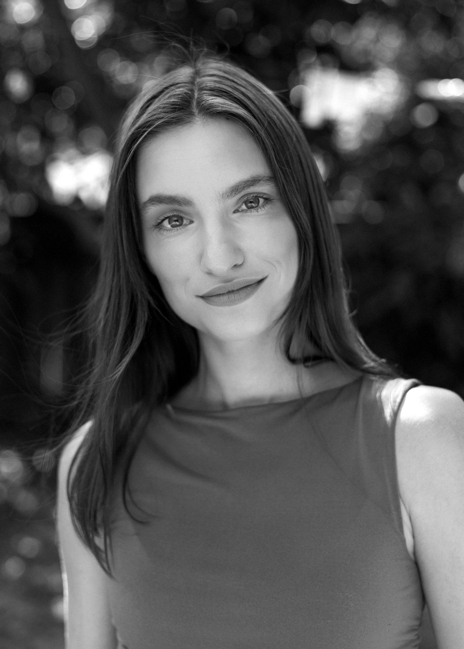 Black and white portrait of a young woman with long dark hair smiling outdoors with blurred foliage background.