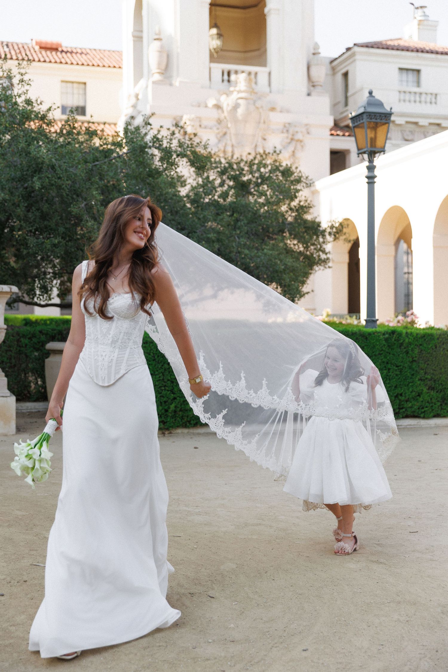Bride in white corset gown holds veil as flower girl walks behind her outdoors near elegant arched building.