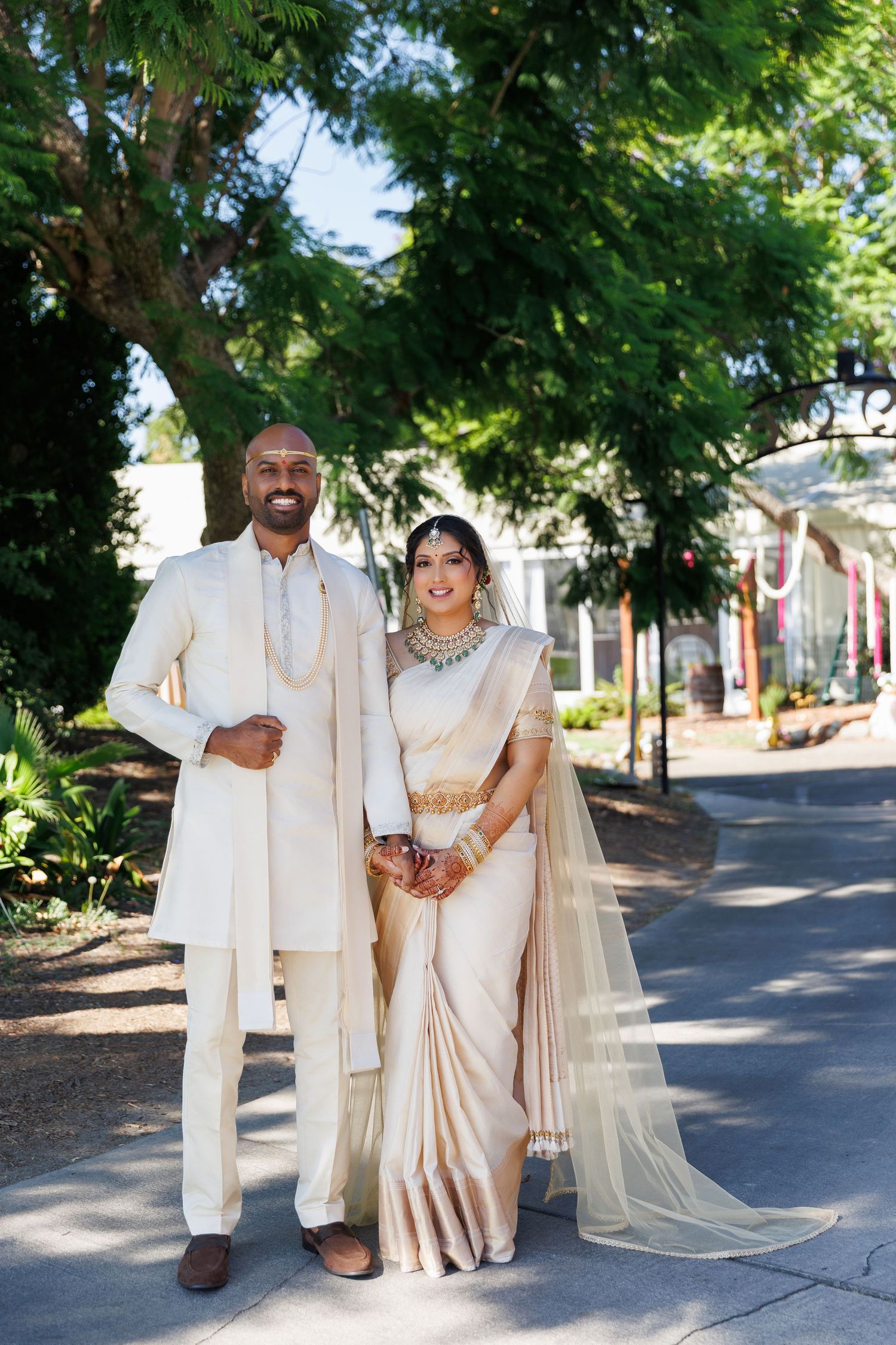 Indian couple in traditional cream wedding attire standing outdoors, bride in saree with gold jewelry and groom in sherwani.