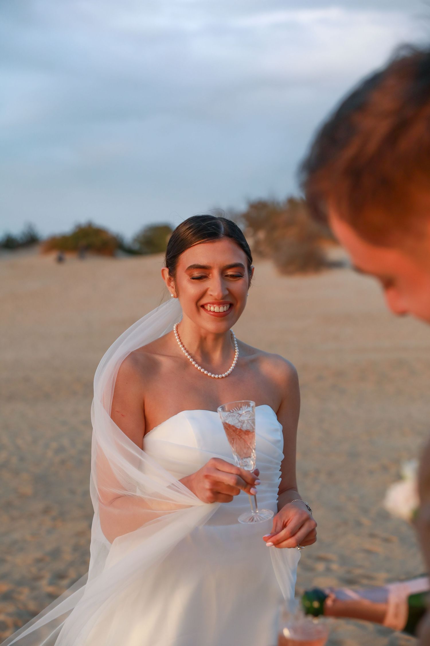 A bride in pearls and veil laughs joyfully during a beach celebration.