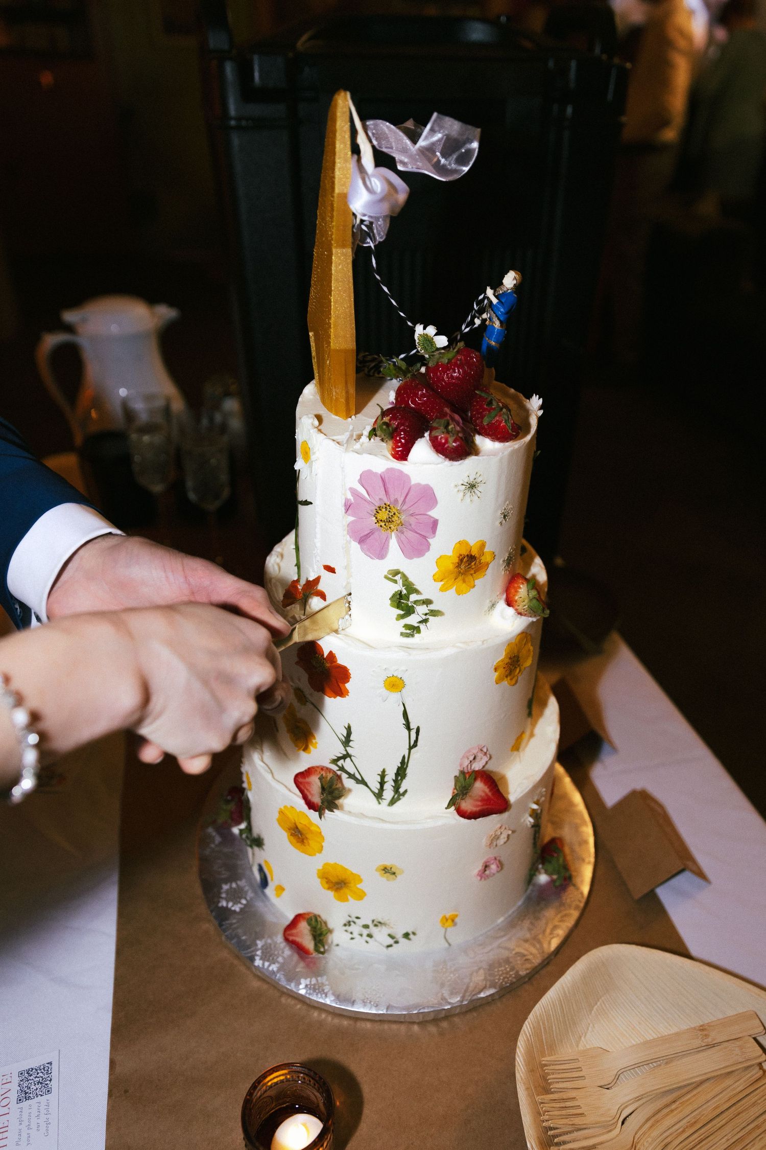 A white wedding cake decorated with colorful flowers and berries being cut at a reception.